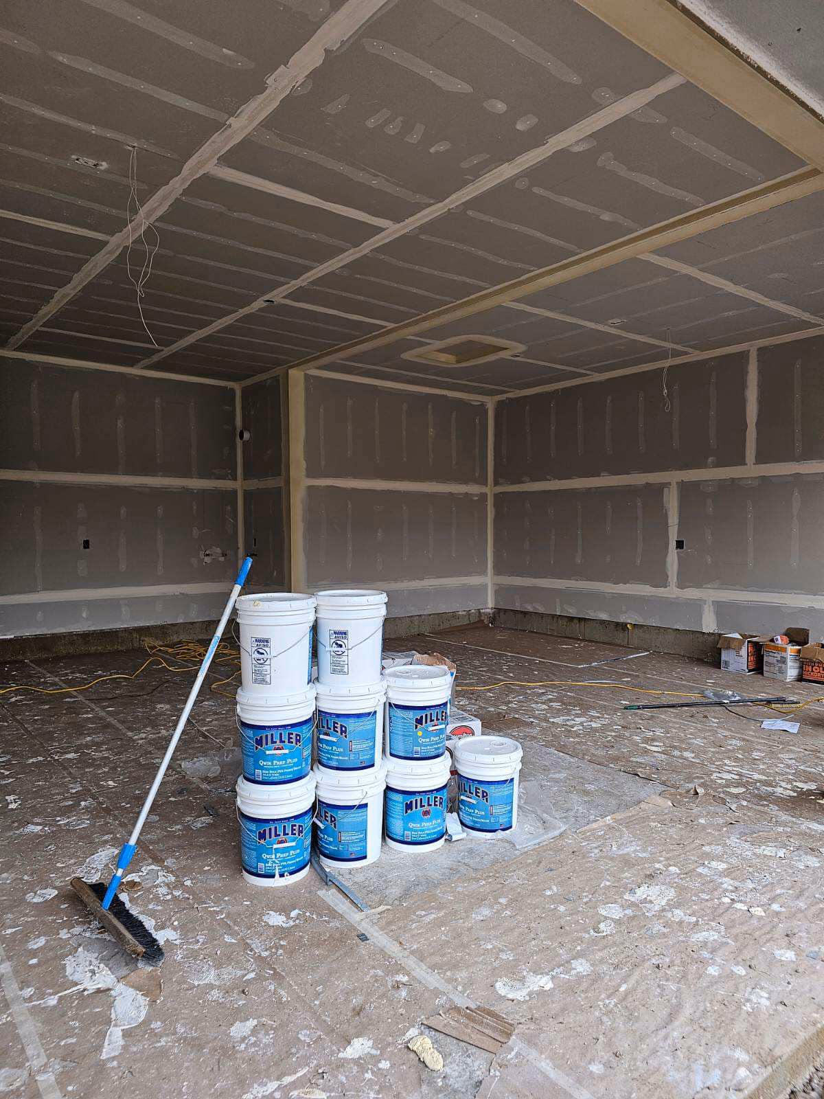 Unfinished room with drywall, buckets of Miller Quick Prep Plus, and a broom on a dusty floor.