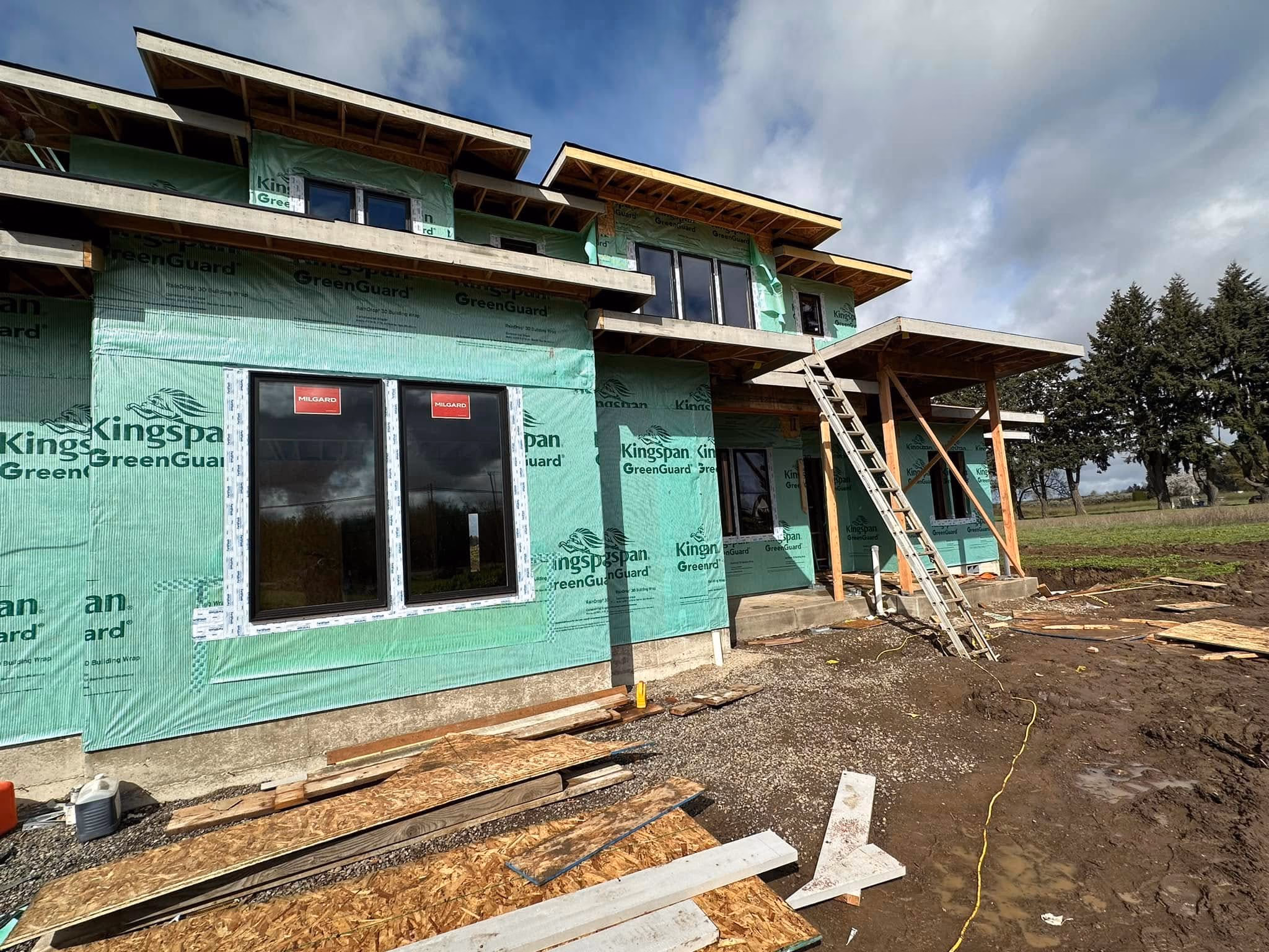 Partially constructed house with green weather-resistant wrap, windows installed, and construction materials nearby.