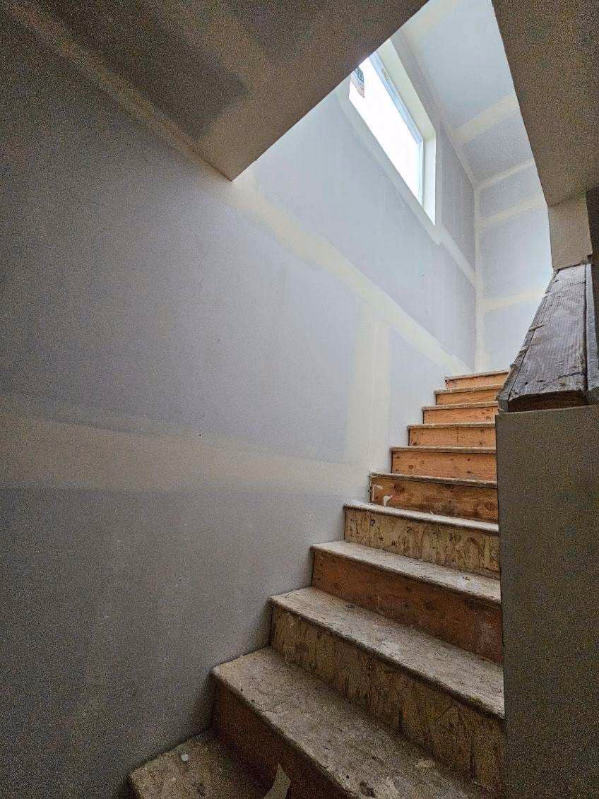 Unfinished wooden stairs leading up to a bright window in a room with drywall walls.