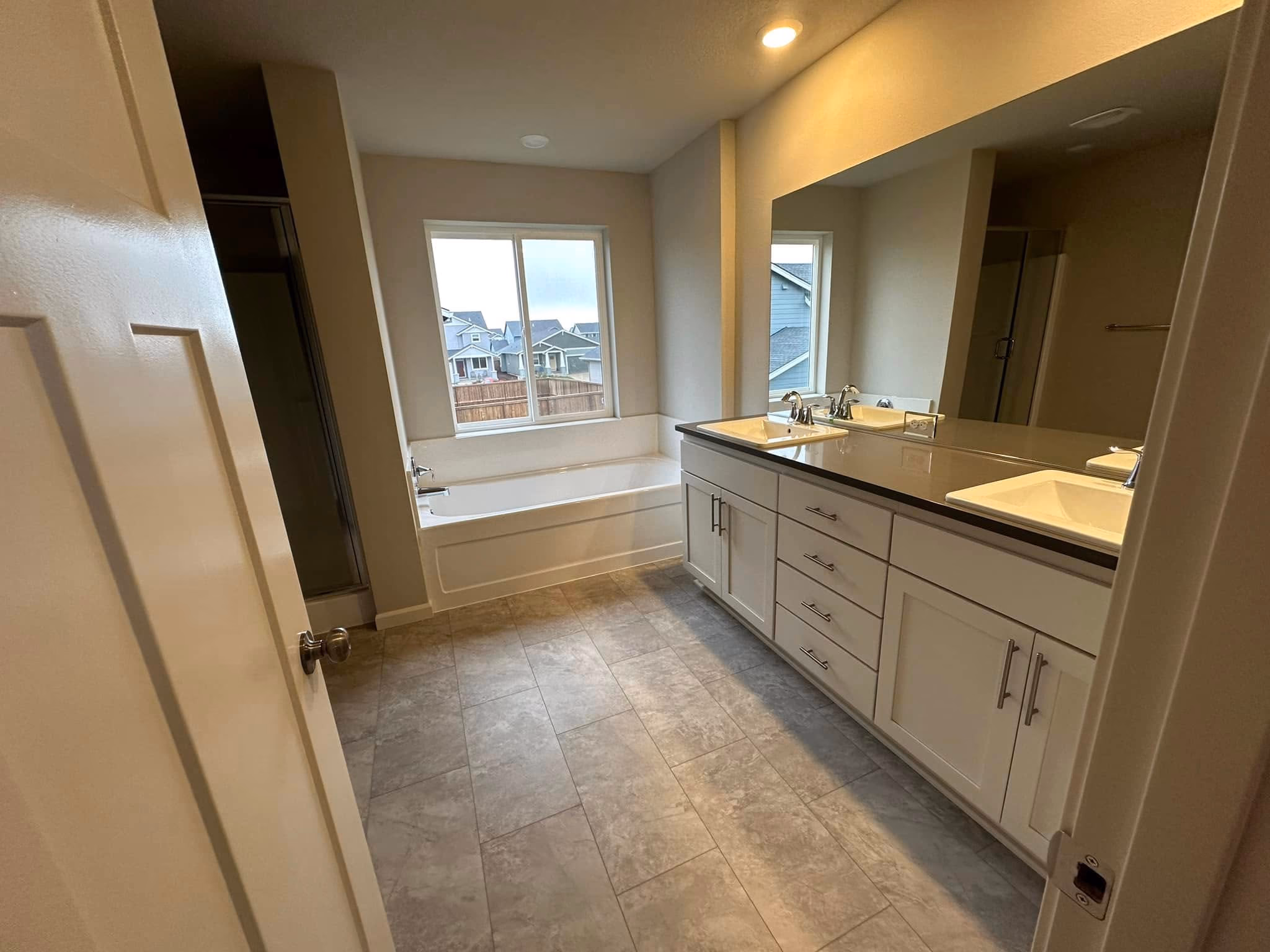 Modern bathroom with white double sinks, large mirror, bathtub below window, and walk-in shower.
