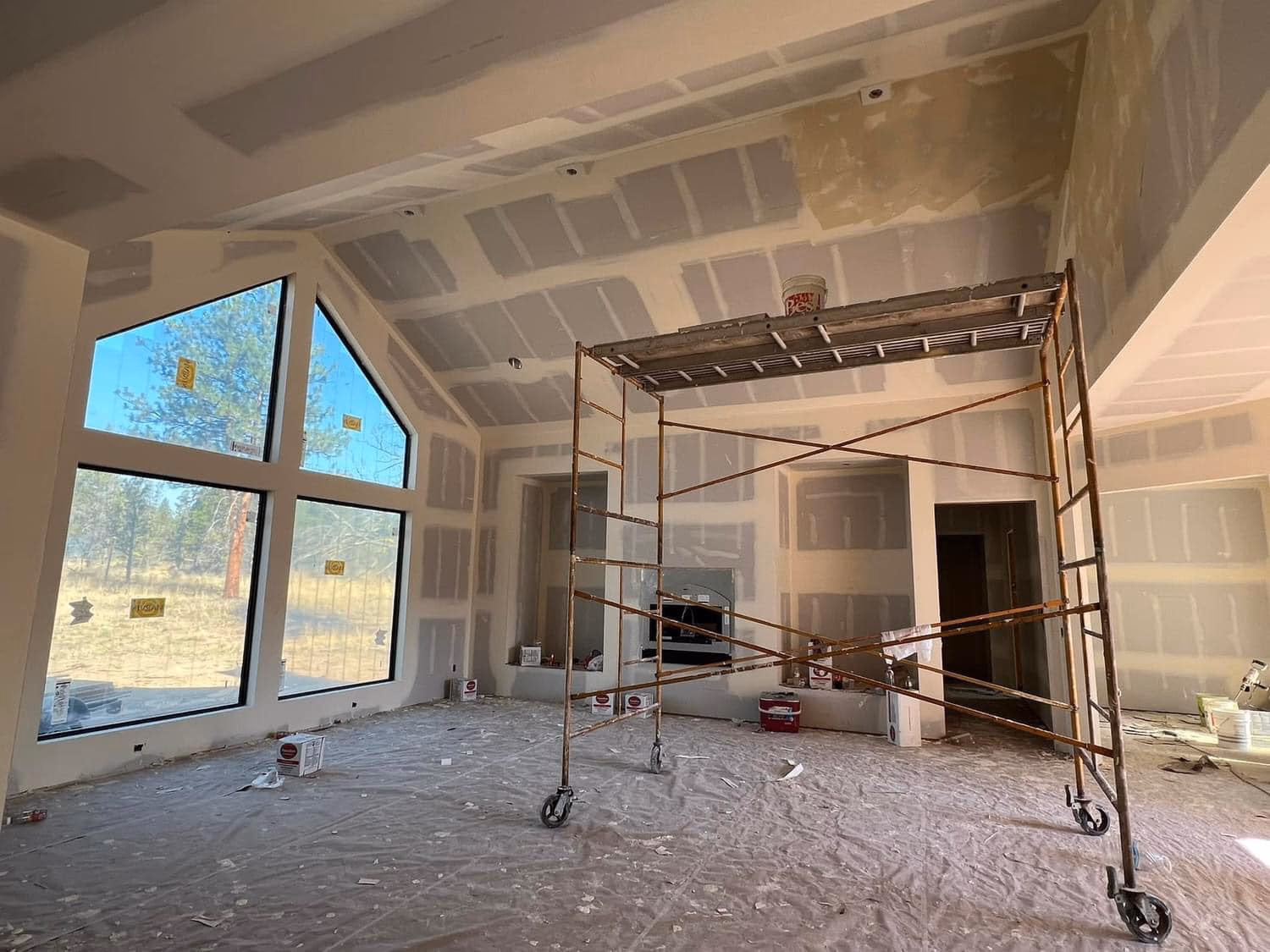 Interior of a room under construction with drywall, large windows, scaffold, and plastic-covered floor.