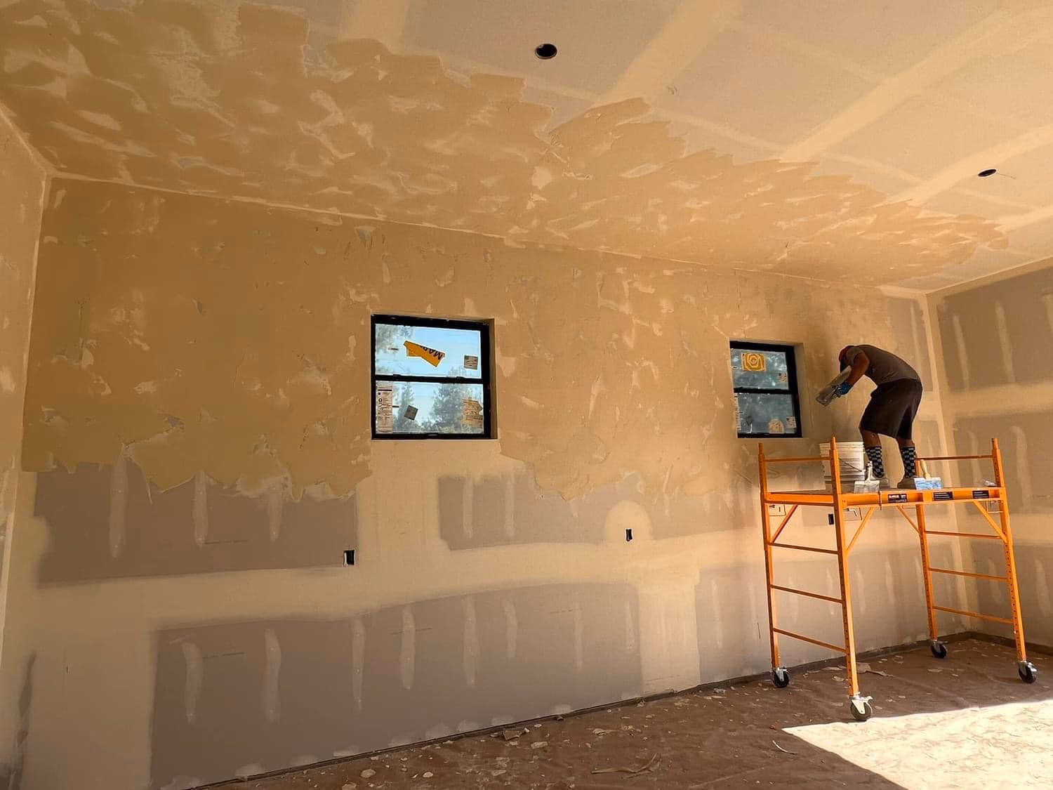 Worker on orange scaffold applying plaster to an unfinished interior wall with two windows.