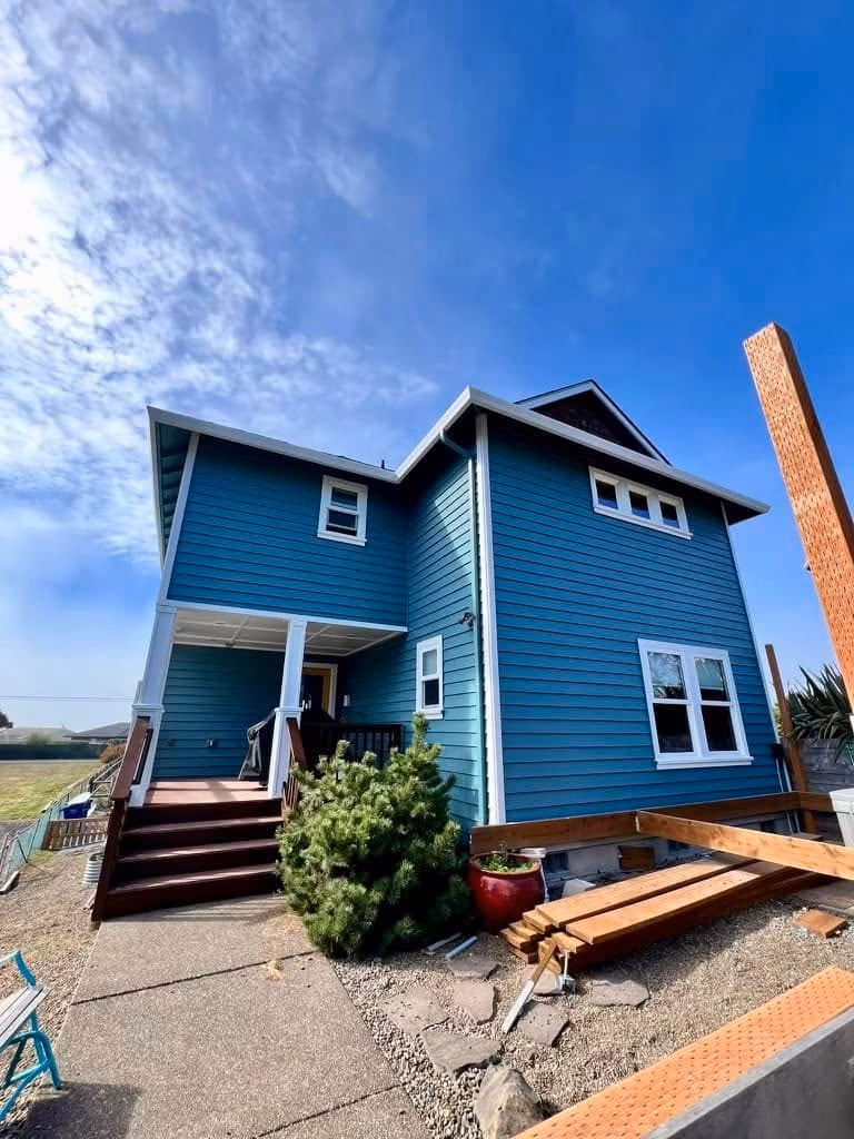 Two-story blue house with white trim, stairs, bush, and wooden beams on gravel yard under a partly cloudy sky.