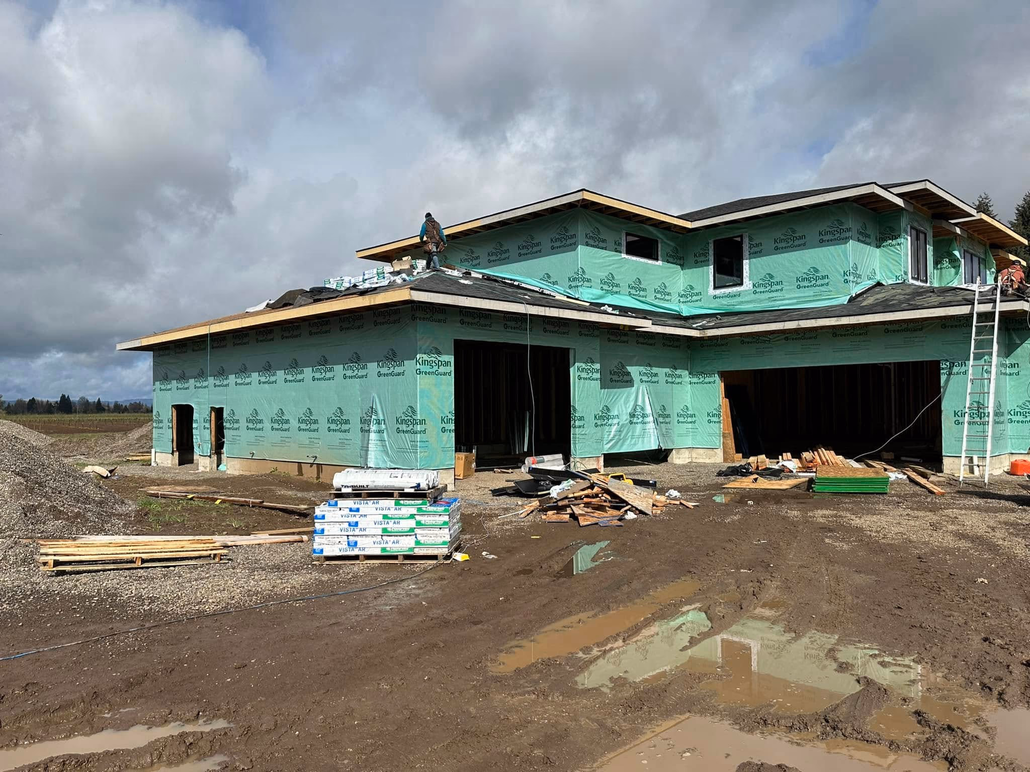 Partially constructed house wrapped in green Kingspan sheathing with workers installing a roof on a muddy site.