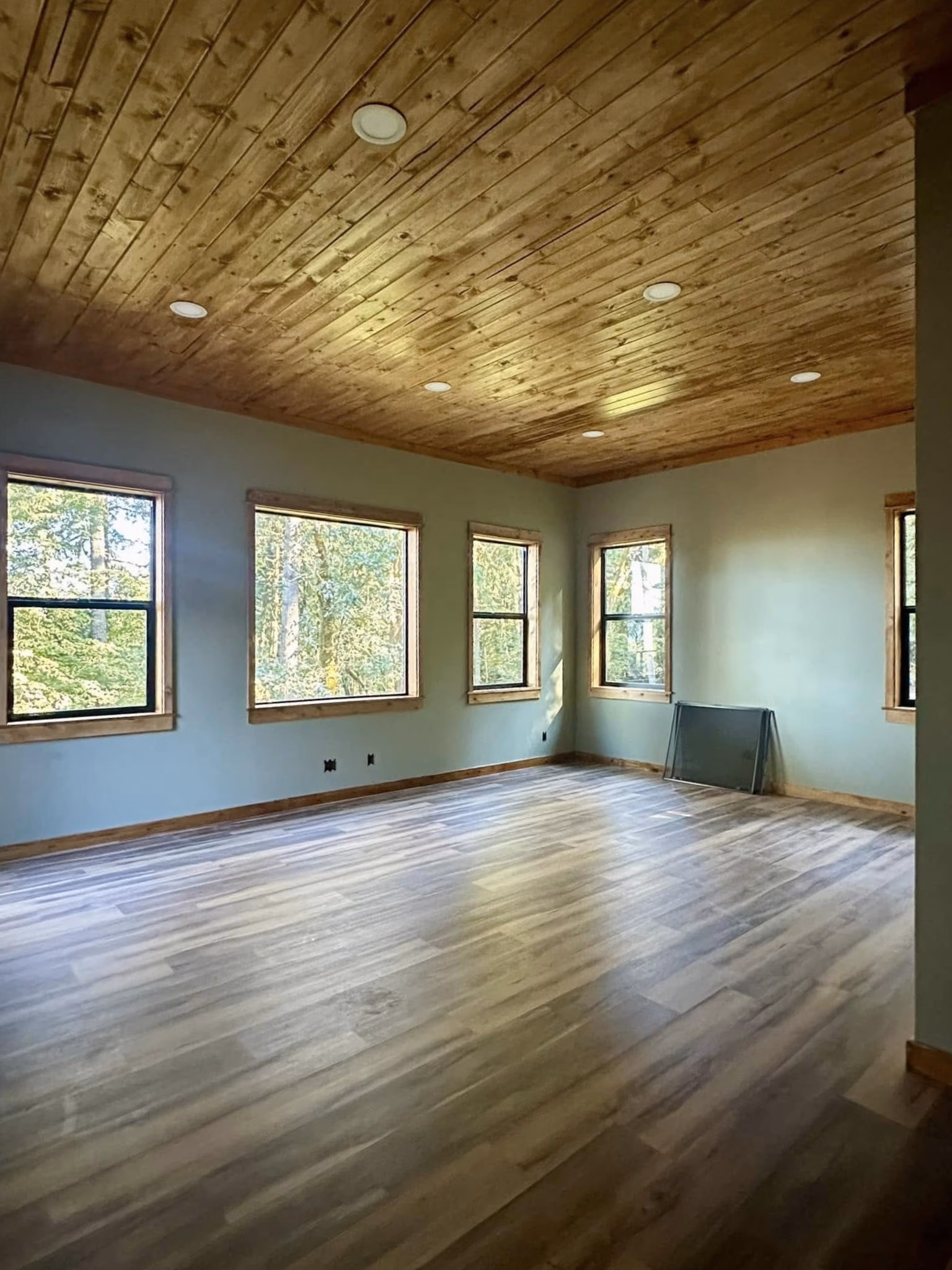 Empty room with wooden ceiling, light wood floor, and several windows showing green trees outside.