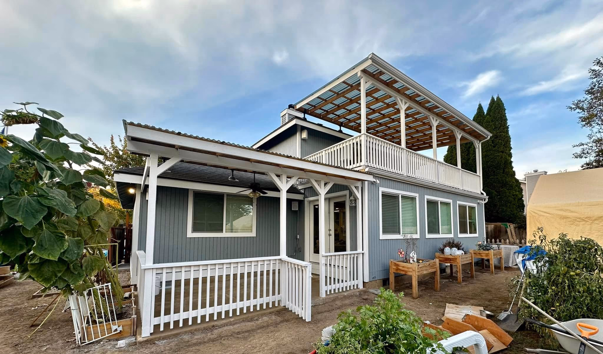 Two-story gray house with white trim, covered porches, garden beds, and green plants in yard.