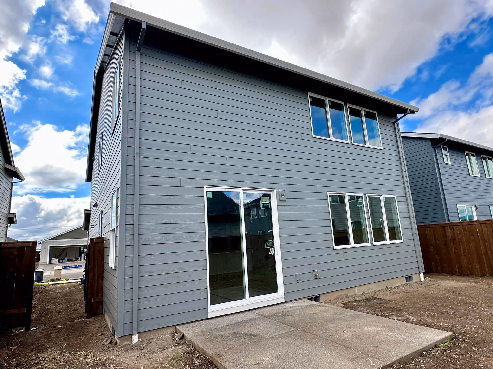 Gray two-story house with sliding glass door and concrete patio under a cloudy blue sky.