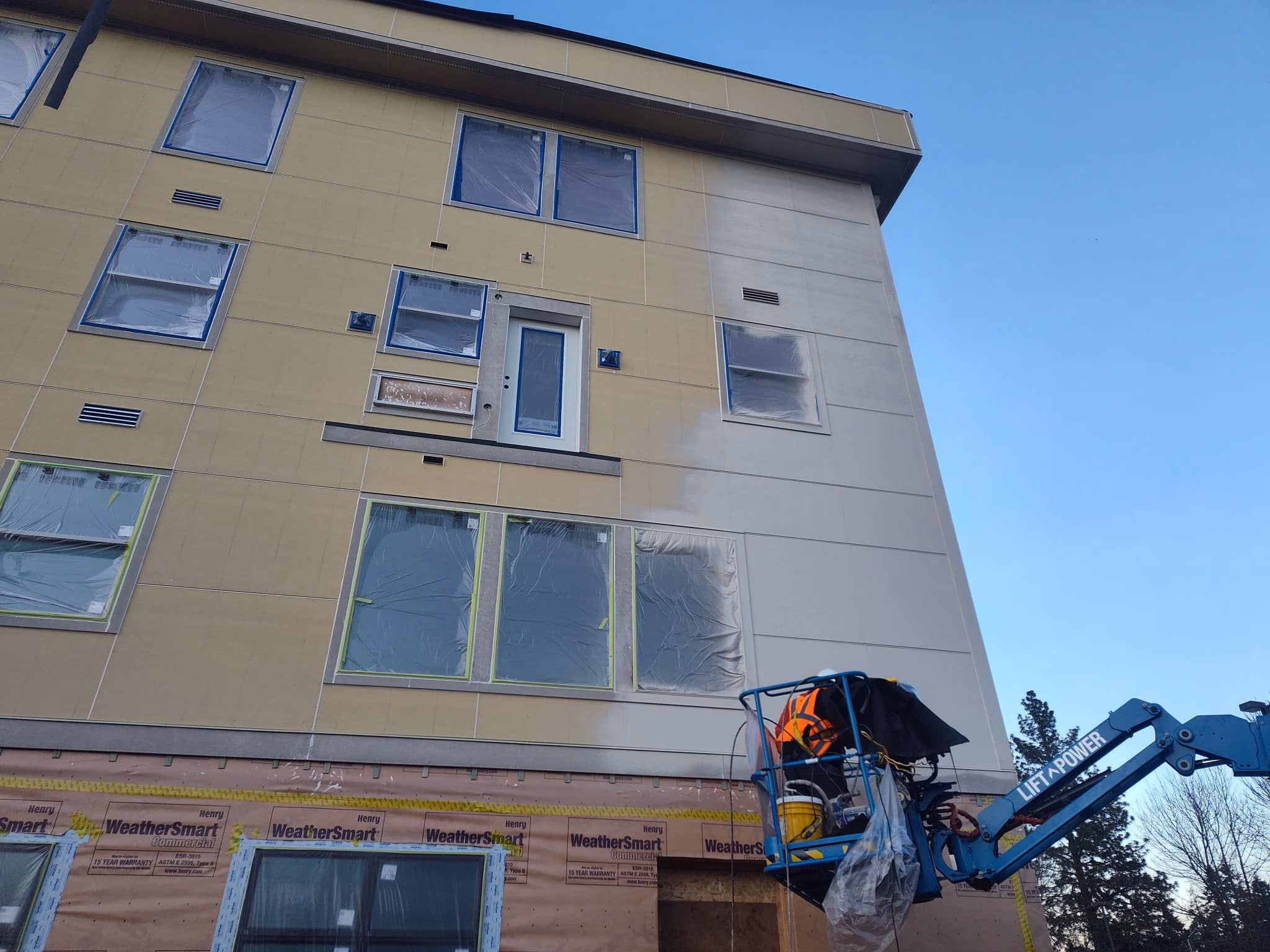Construction worker on lift painting beige exterior wall of multi-story building with covered windows.