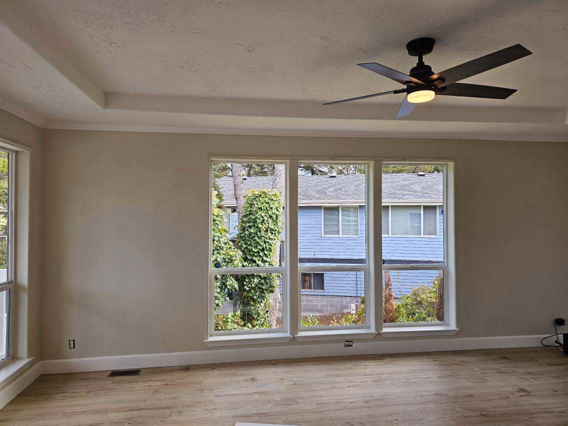 Empty room with beige walls, wooden floor, large triple window, and ceiling fan with light.