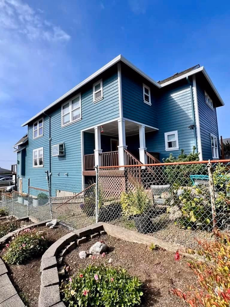 Blue two-story house with white trim, a fenced yard, and garden beds with flowers and shrubs.