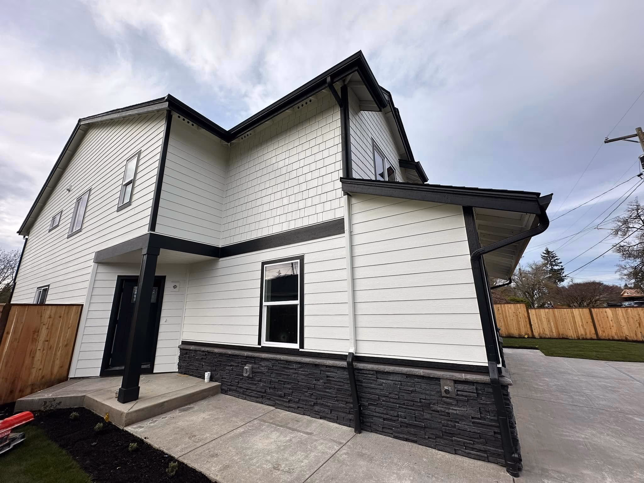 Two-story white house with black trim, stone base, a small porch, and a wooden fence under a cloudy sky.