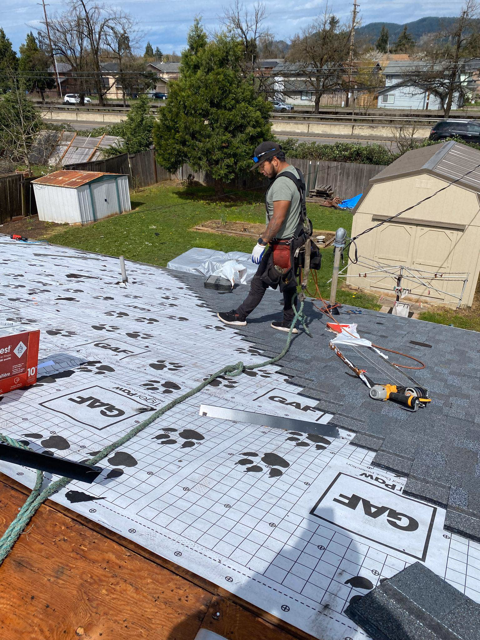 Roofer wearing safety harness installing shingles on a house roof under a partly cloudy sky.