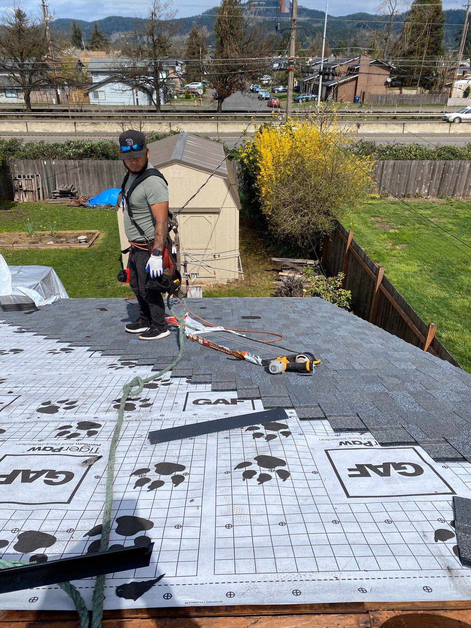 Worker wearing harness installing gray roof shingles on a residential rooftop with tools nearby.