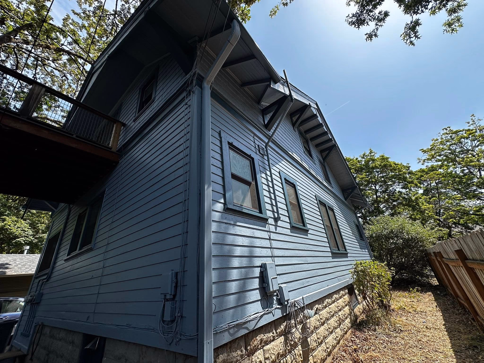 Blue two-story house exterior with multiple windows, electrical boxes, and a wooden fence beside it.