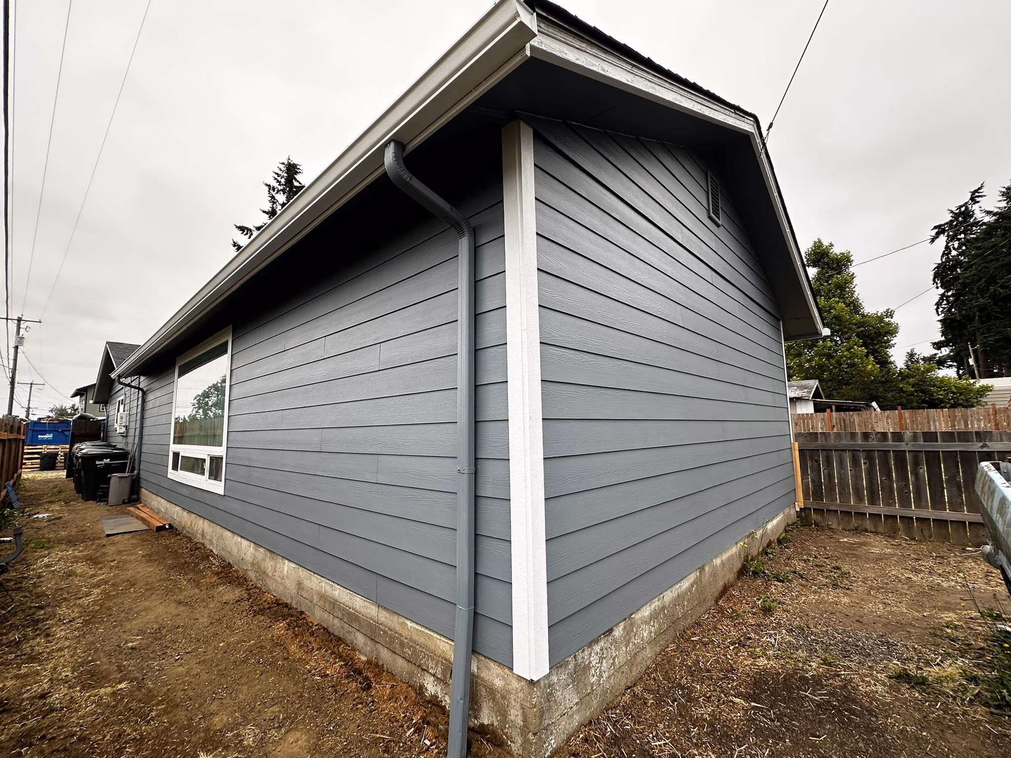 Corner view of a gray house with horizontal siding, a window, gutter, and bare ground around it.