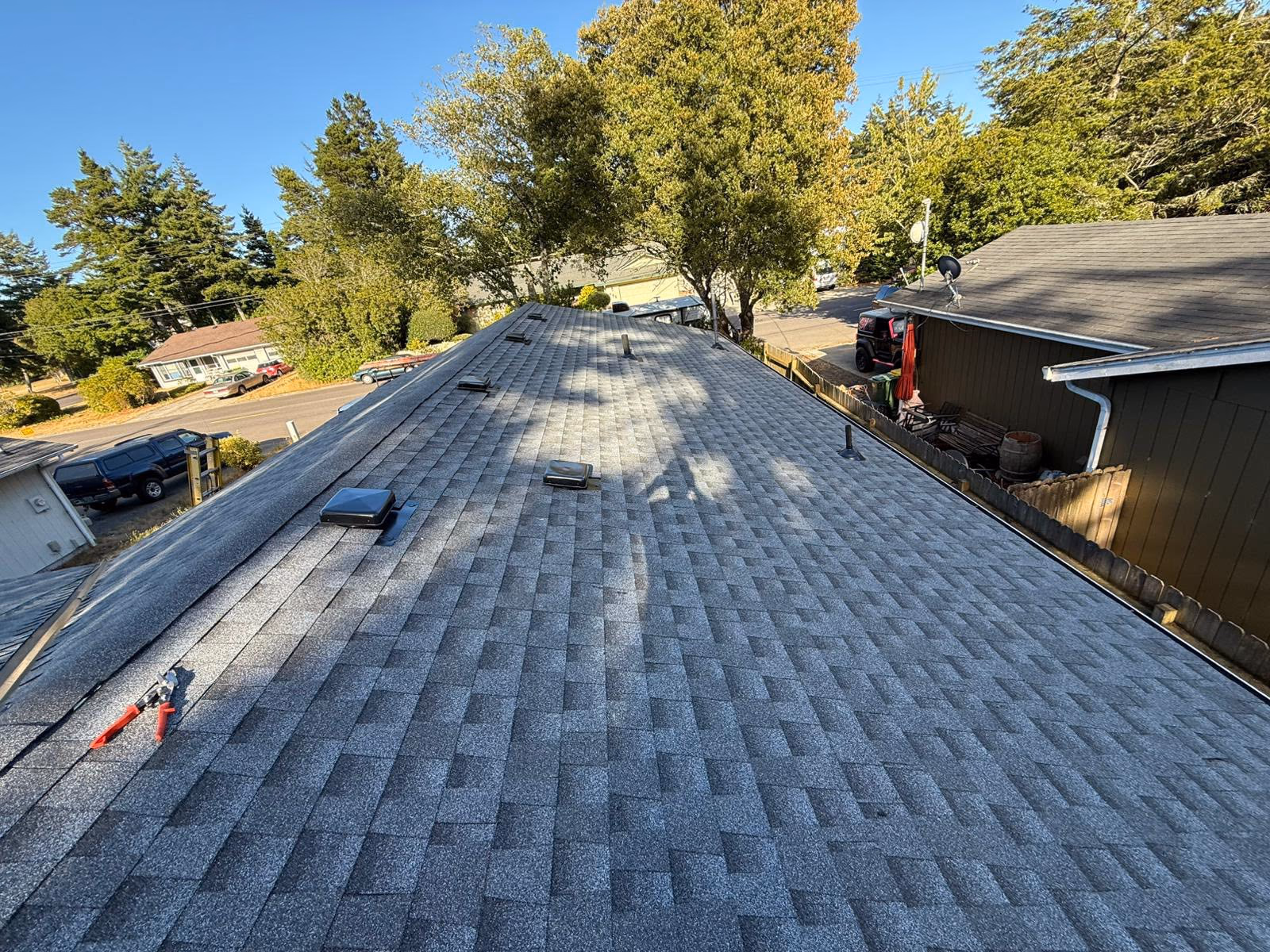 Shingled roof with vent covers and a tool laying near the edge, surrounded by trees and neighboring houses.