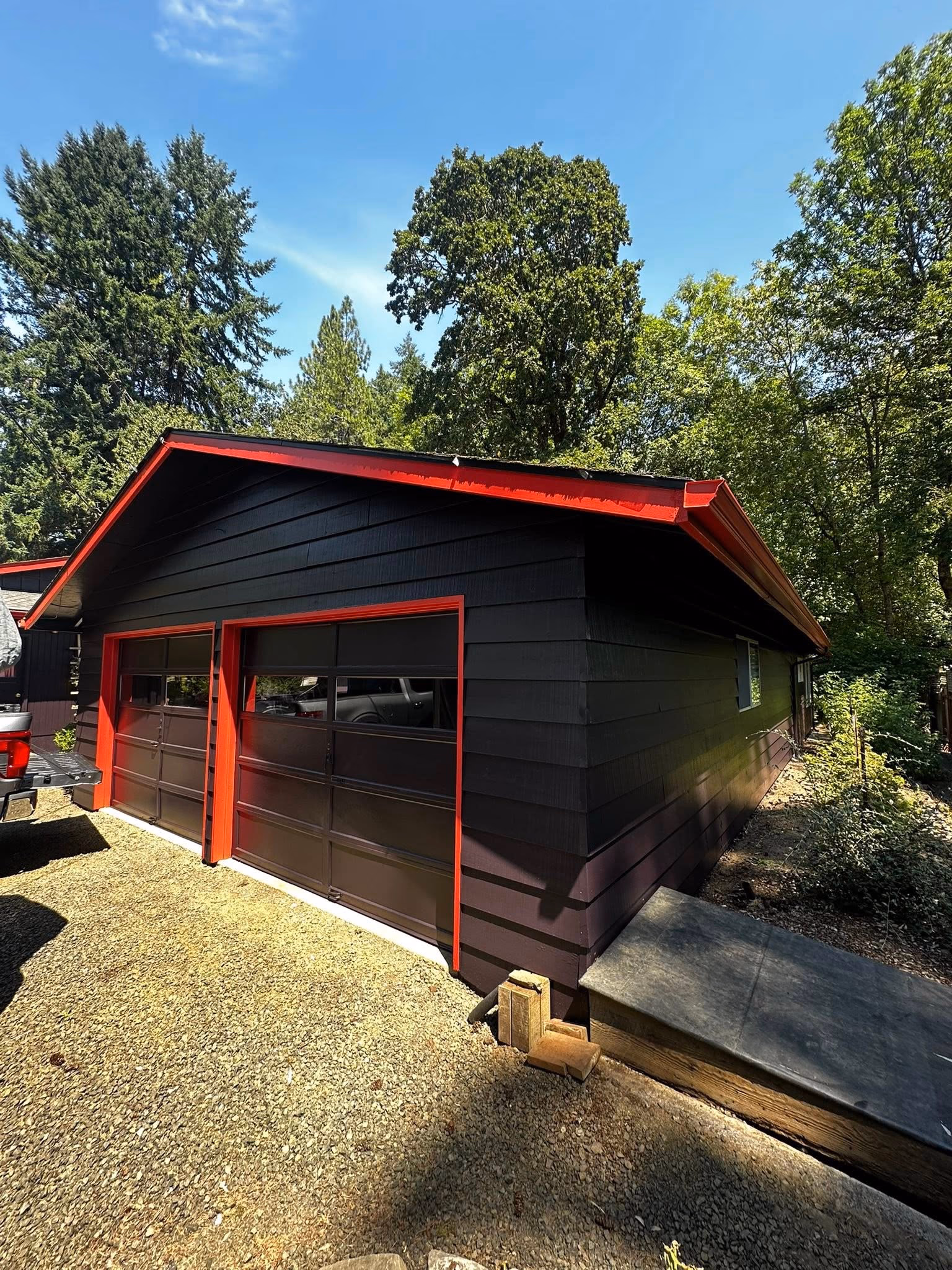 Black garage with red trim surrounded by gravel and green trees under a clear blue sky.