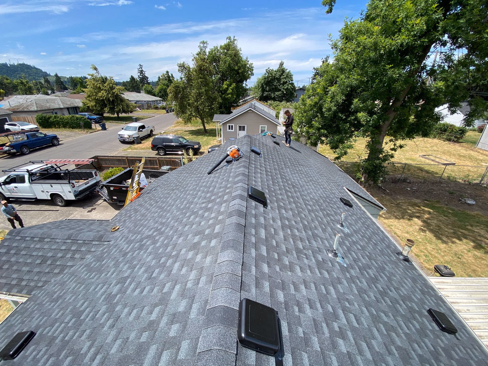 Newly installed gray asphalt shingle roof with vents and worker on a sunny suburban street.