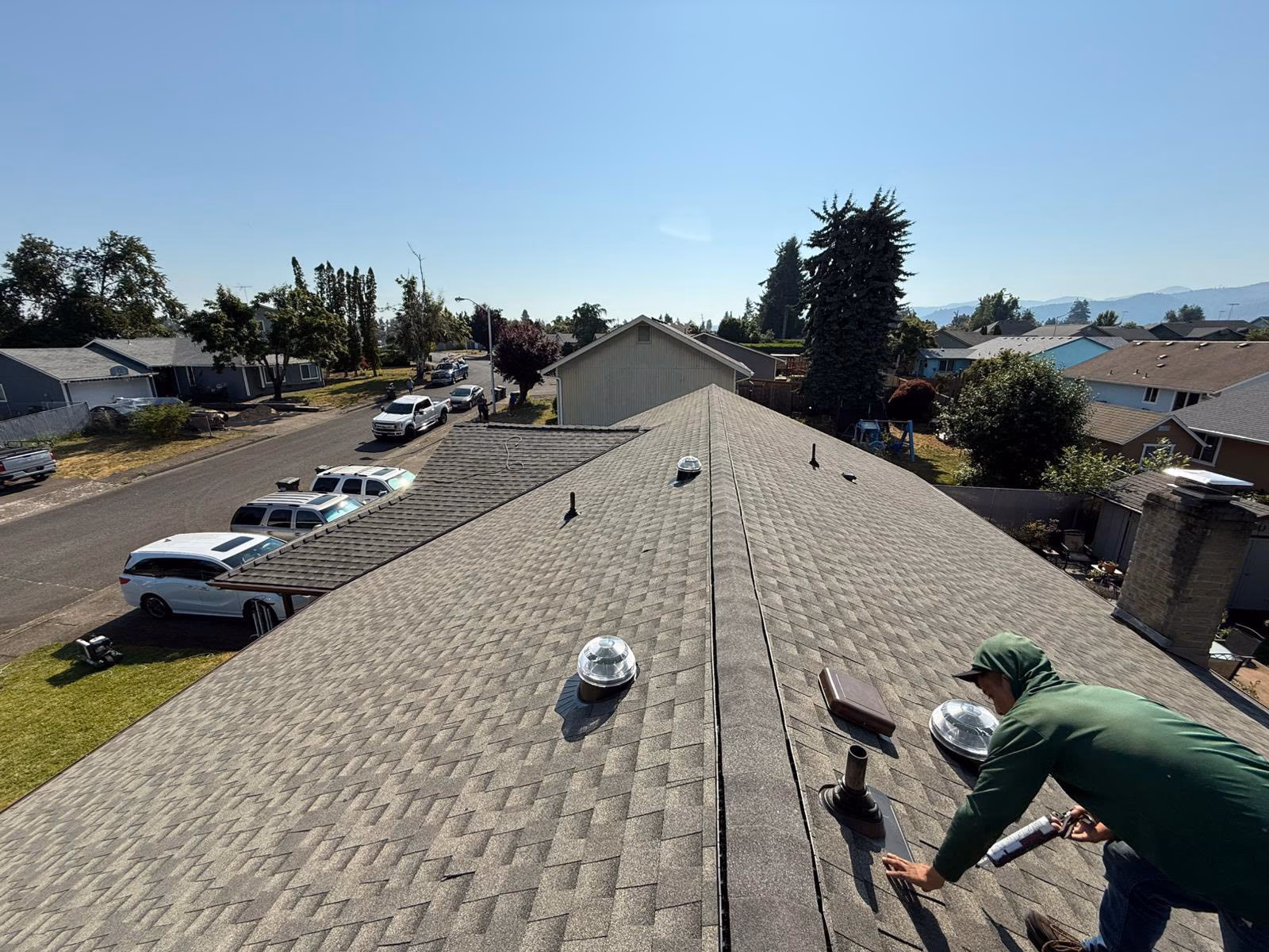 Man in green hoodie working on a residential roof with asphalt shingles on a sunny day.