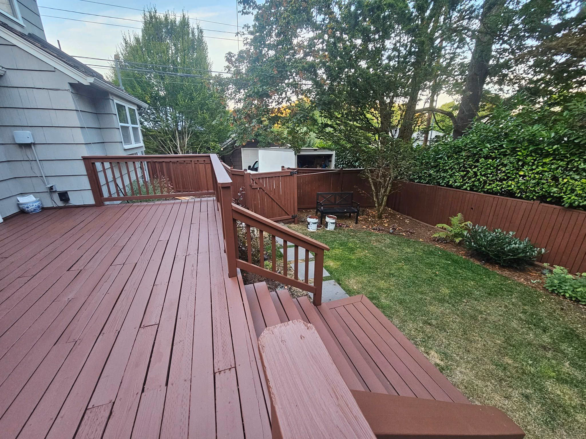 Backyard with brown wooden deck, stairs, lawn, trees, plants, and a small black bench by a fence.