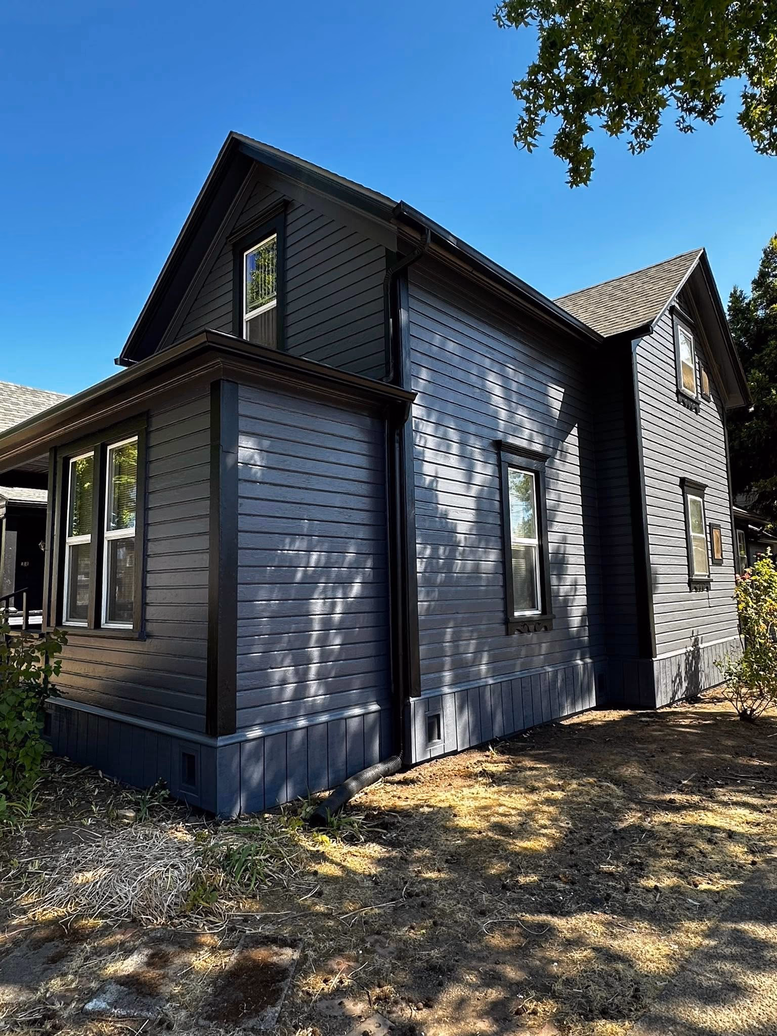 Blue-gray wooden house with black trim under clear blue sky and tree shadows on the facade.