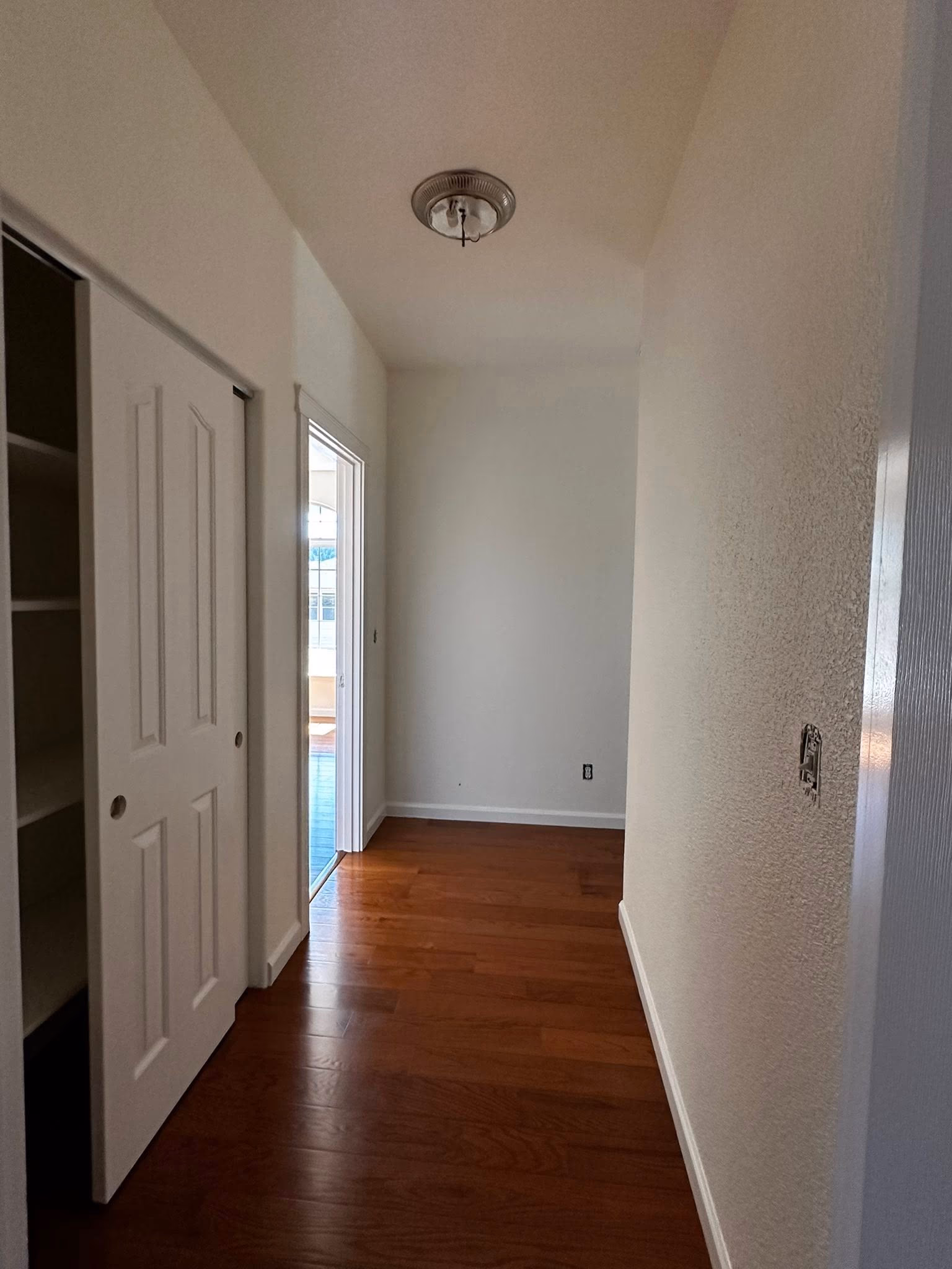 Empty hallway with white walls, wood floor, sliding closet doors, and doorway to sunlit room.