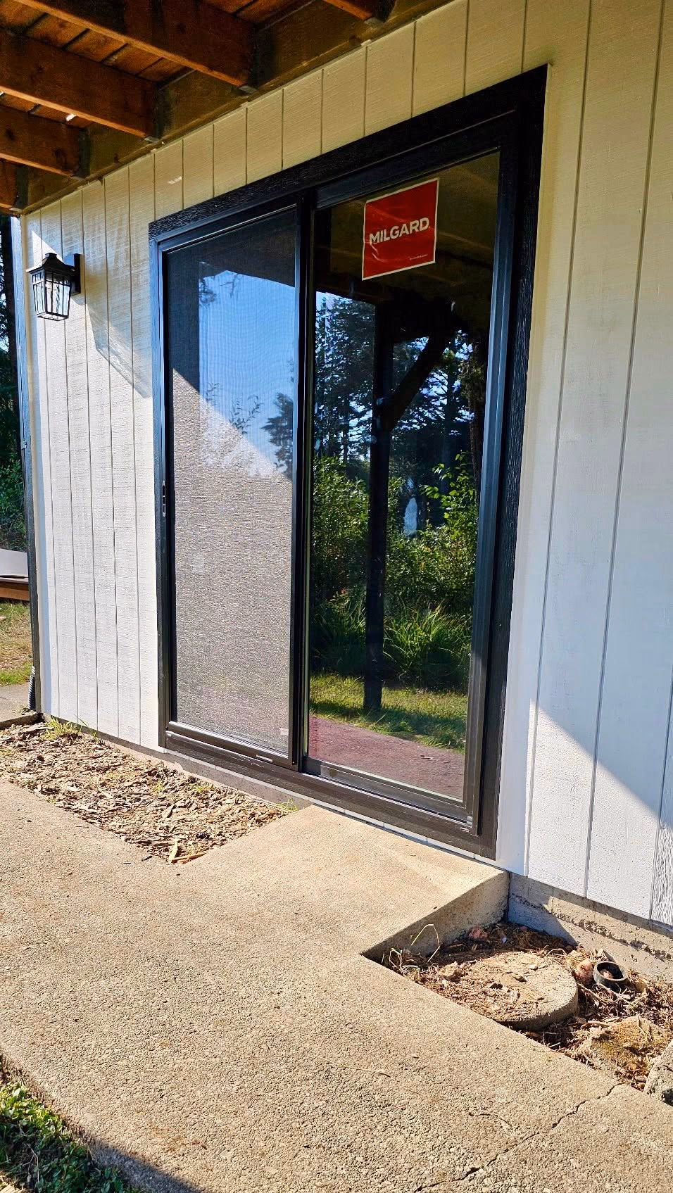 Black-framed sliding glass door with screen on a white paneled house exterior under wooden roof.