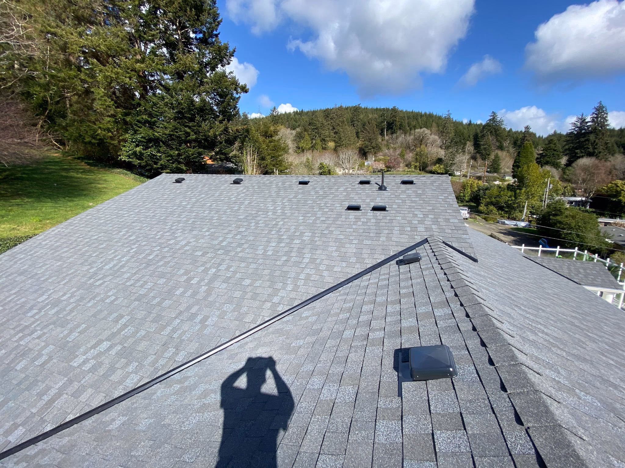 Gray shingled house roof with vents and trees on a sunny day under blue sky with clouds.