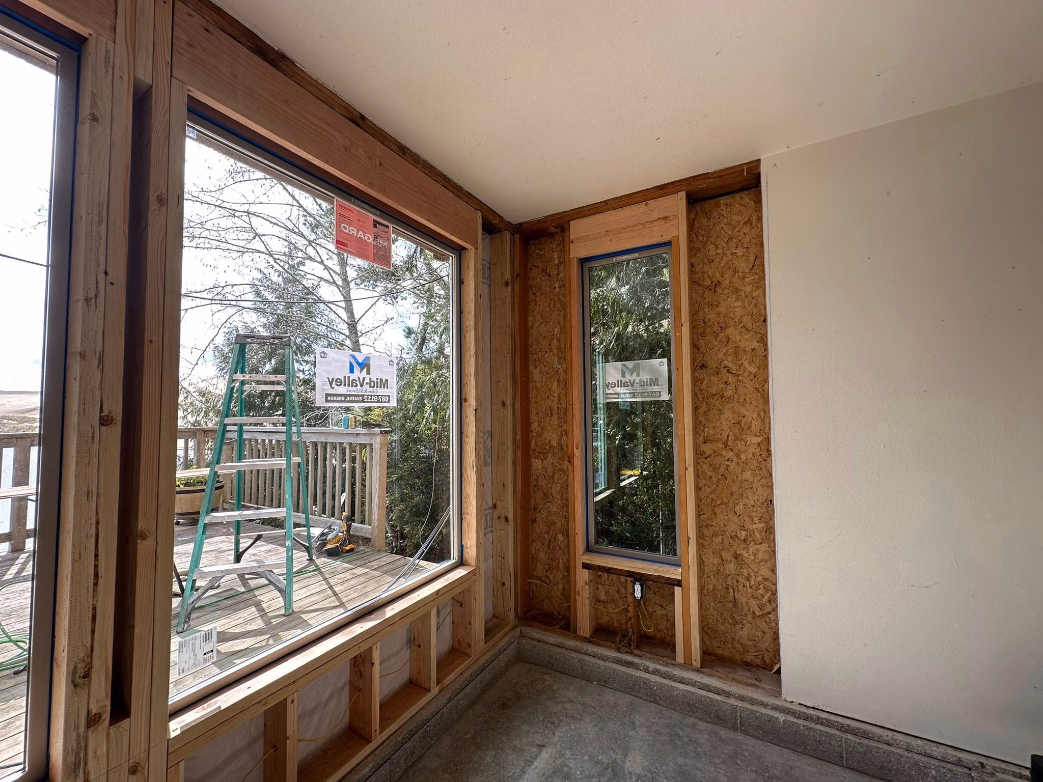 Interior corner of unfinished room with two large windows overlooking a wooden deck and trees.