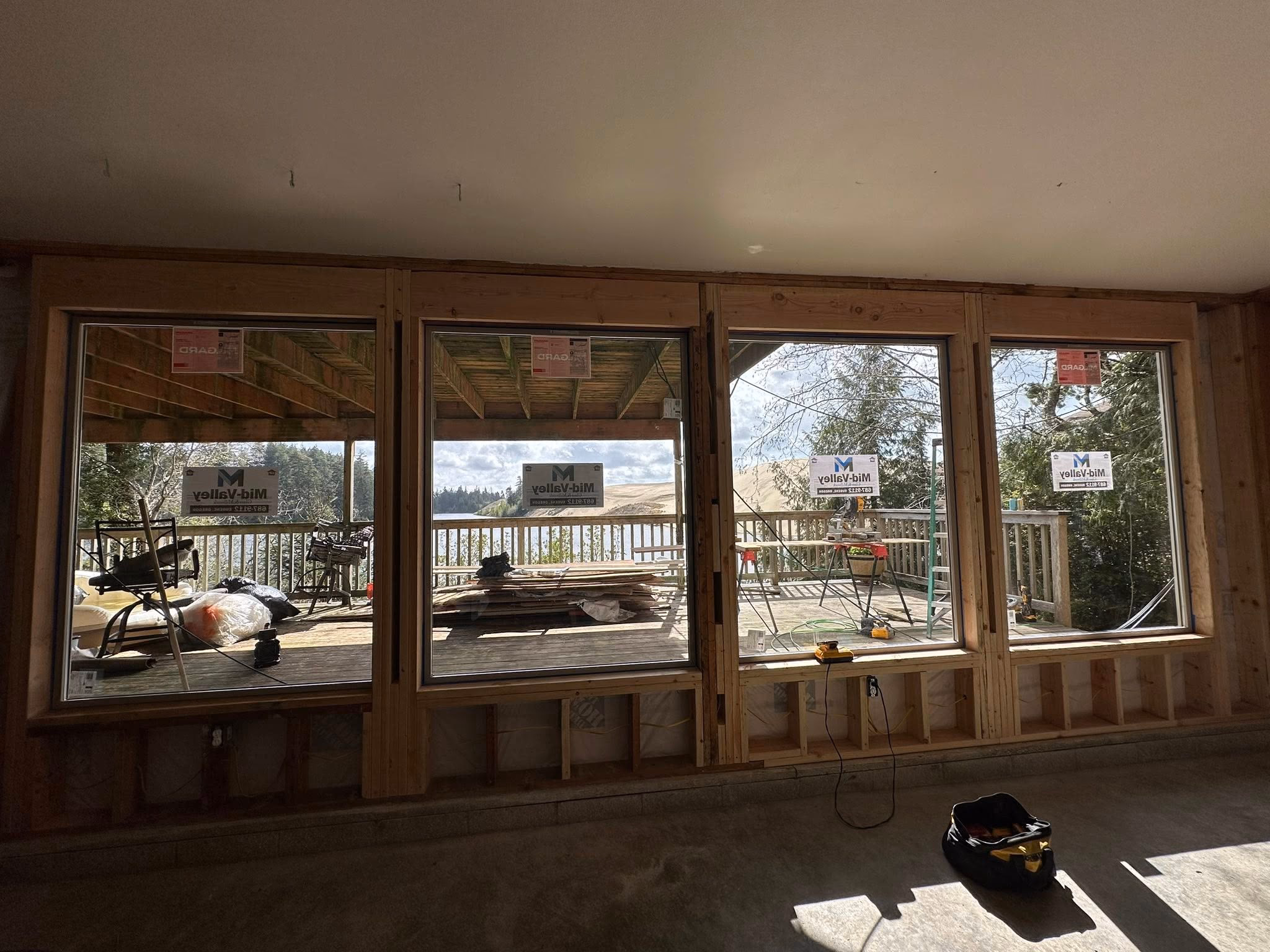 Four large windows under construction show an outdoor deck with tools, wood stacks, and trees beyond.