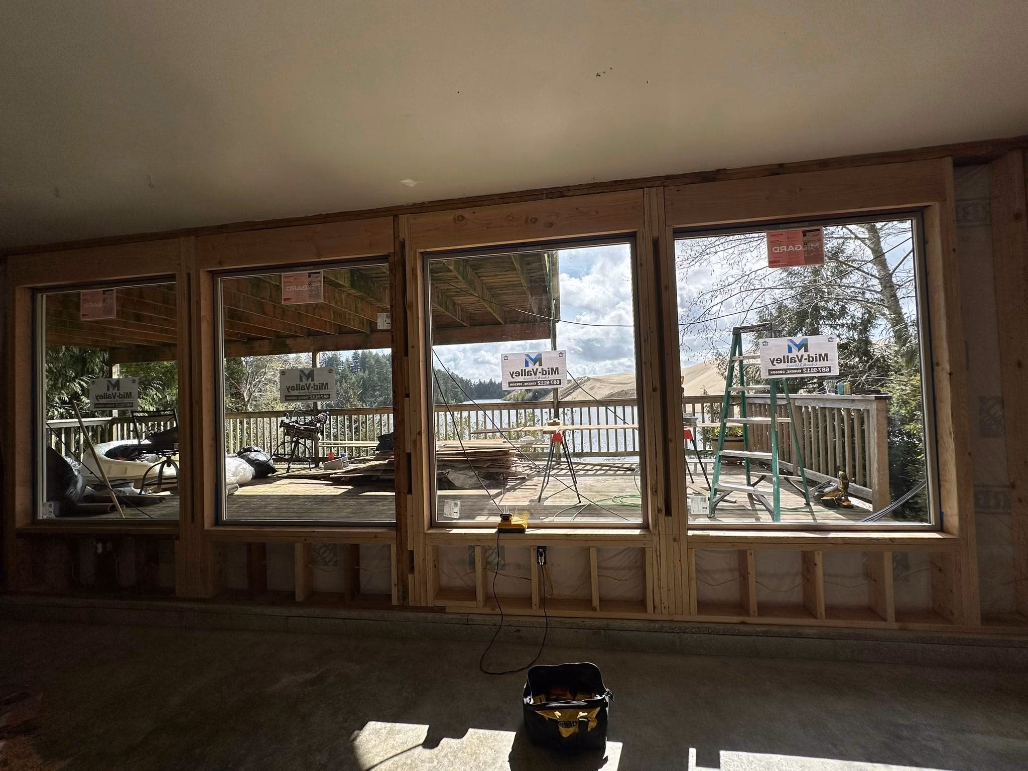 Four large framed windows under construction showing a deck with construction tools and a lake view outside.