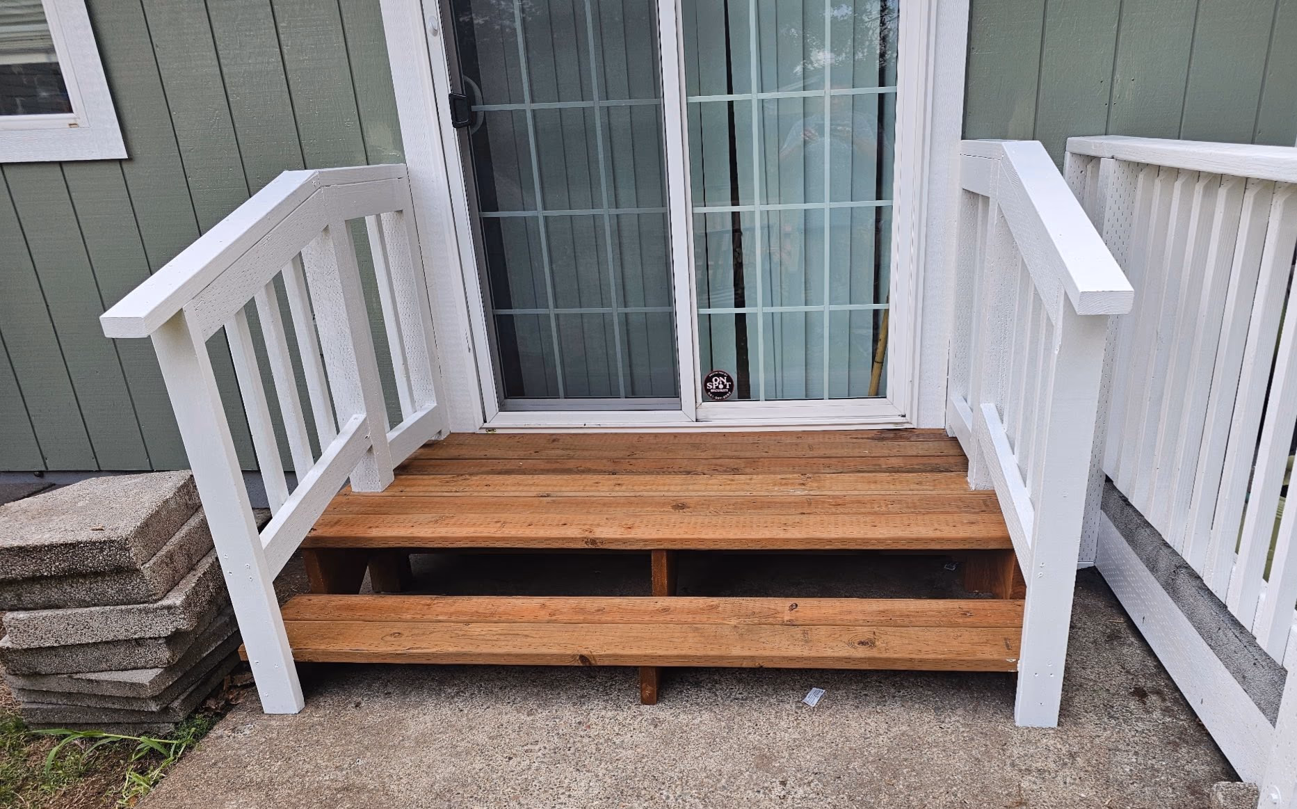 Small wooden porch with two steps and white railings in front of a sliding glass door.