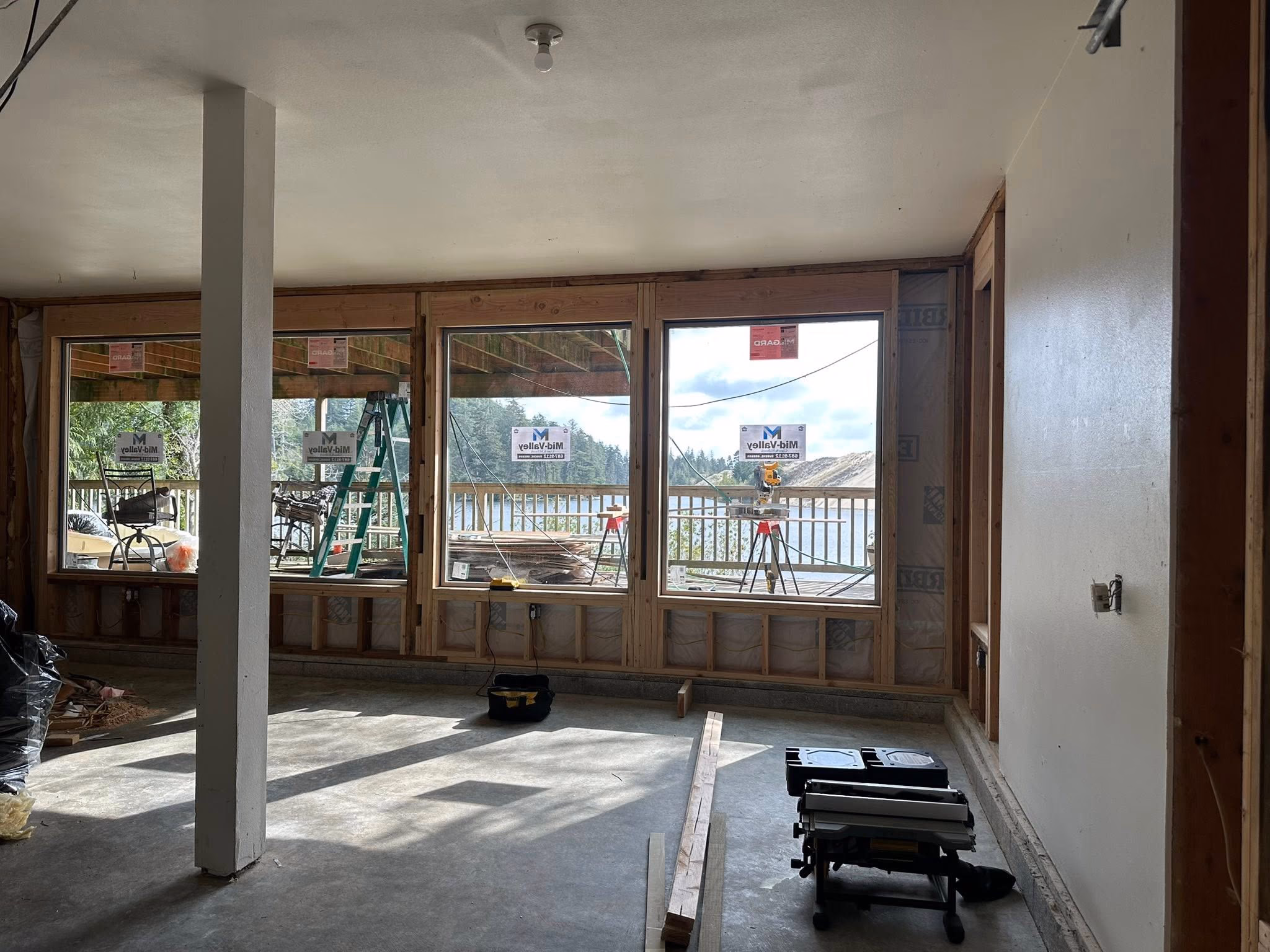 Room under construction with large windows showing an outdoor deck, ladder, and lake view.