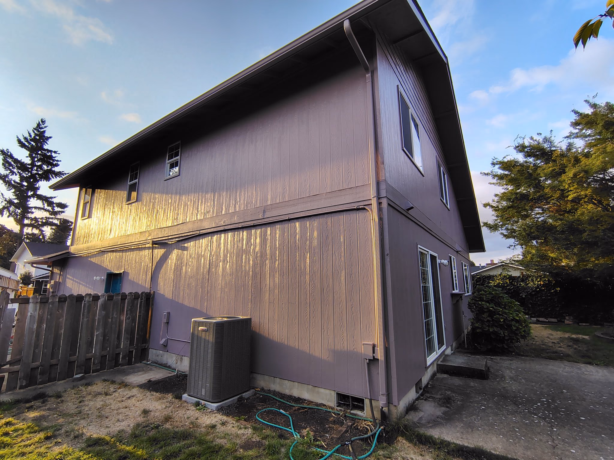 Two-story purple house exterior with windows, air conditioner, and wooden fence in yard during sunset.