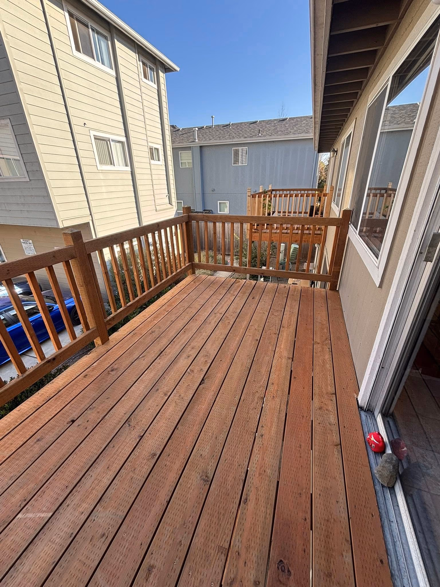 Empty wooden balcony with railing attached to a building, overlooking neighboring houses under a clear sky.