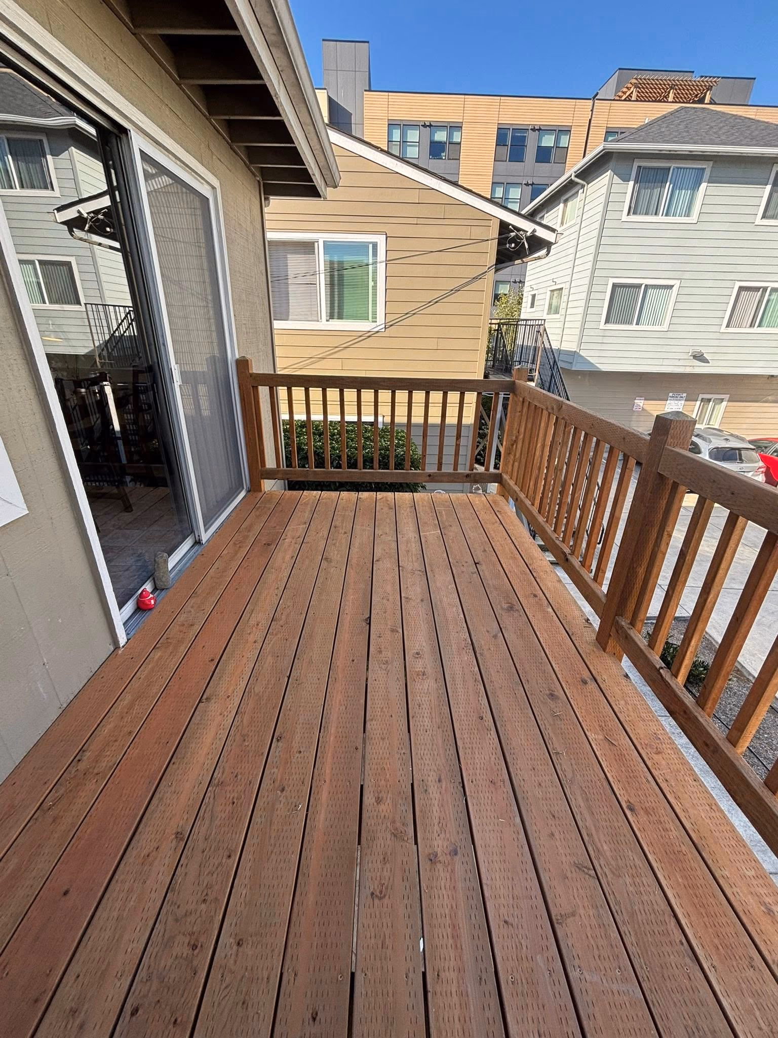 Wooden balcony with railing attached to a house, overlooking neighboring buildings and parked cars.
