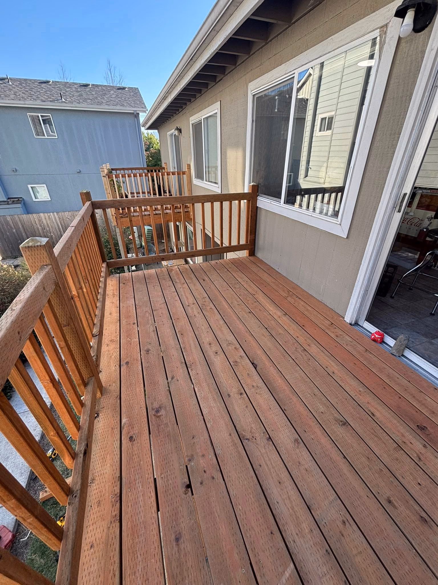 Wooden upper deck attached to house with railing, sliding door, and neighboring blue house in background.
