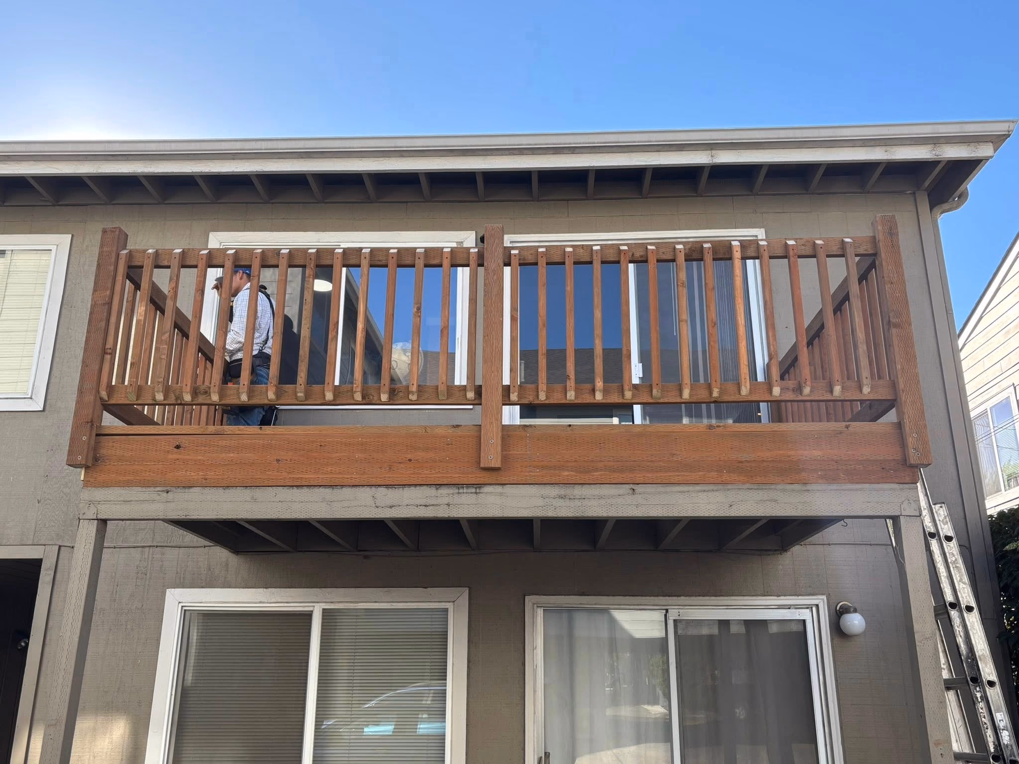 Person working on a wooden balcony railing attached to a two-story beige house under blue sky.