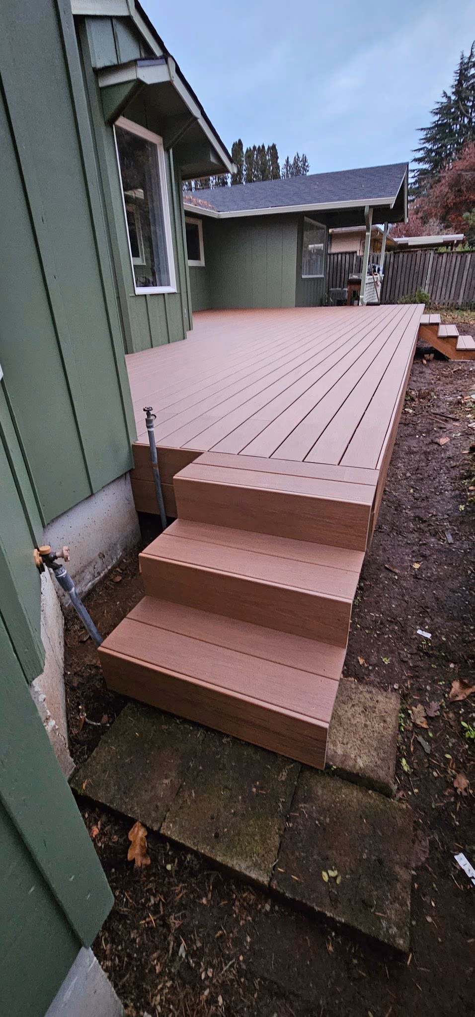 Newly built brown wood composite deck with steps attached to a green house exterior.