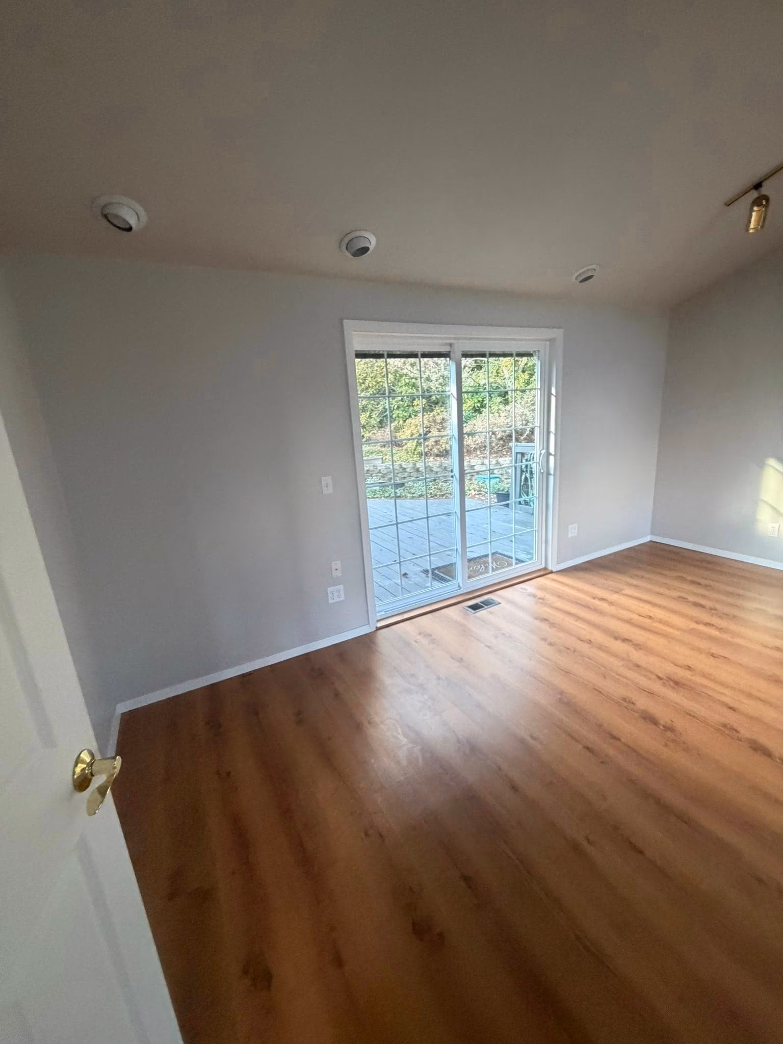 Empty room with wooden floor, sliding glass door, and light gray walls, leading to outdoor deck.