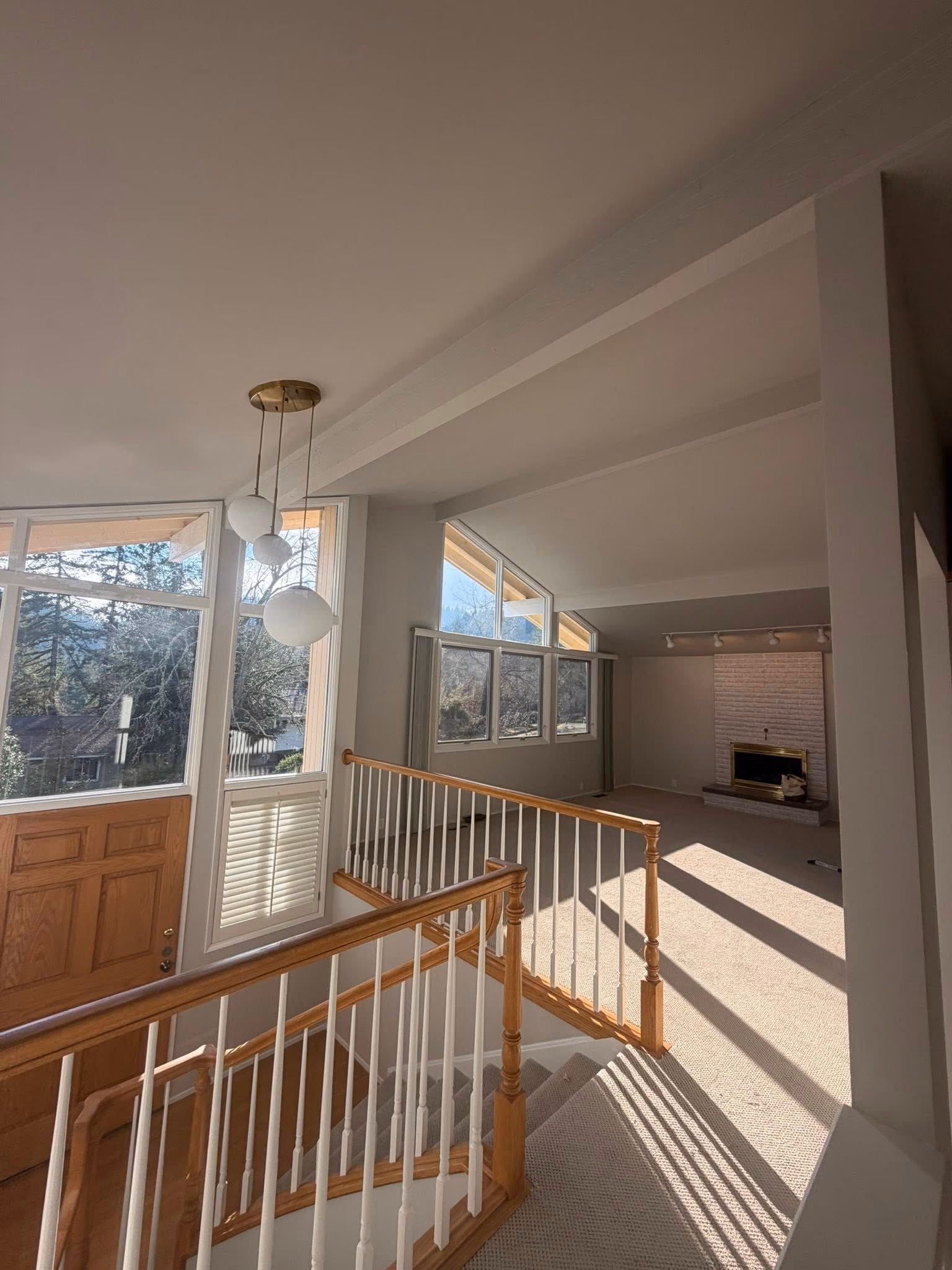 Interior stairway with wooden railing leading to sunlit living room with fireplace and large windows.