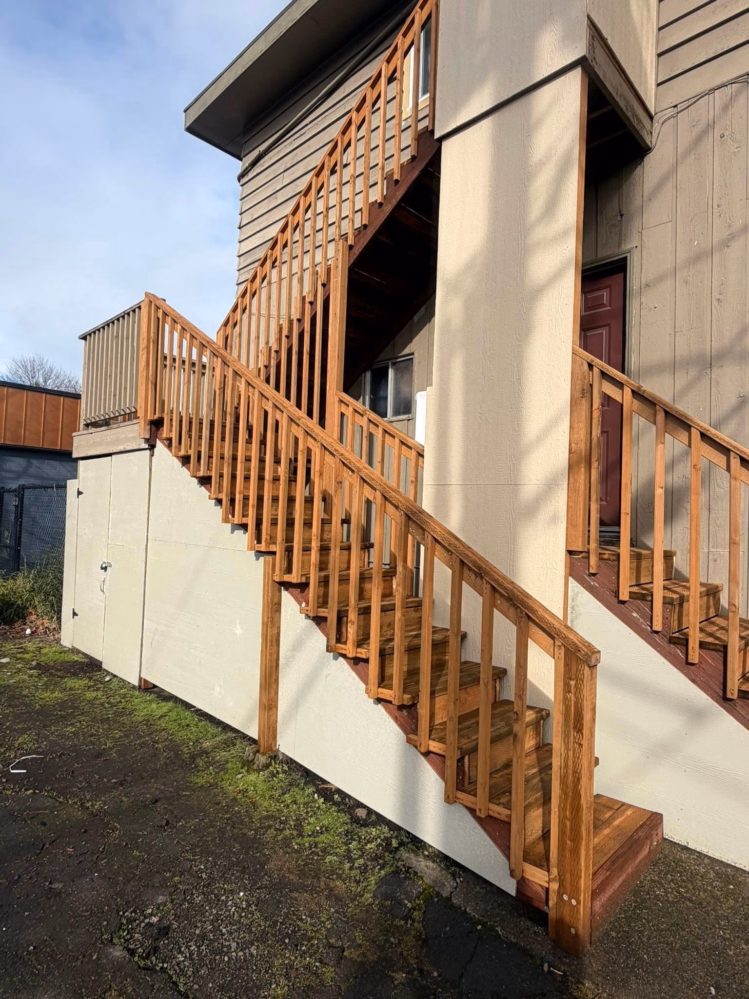 Outdoor wooden staircase with railings leading to two levels of a beige building with a red door.