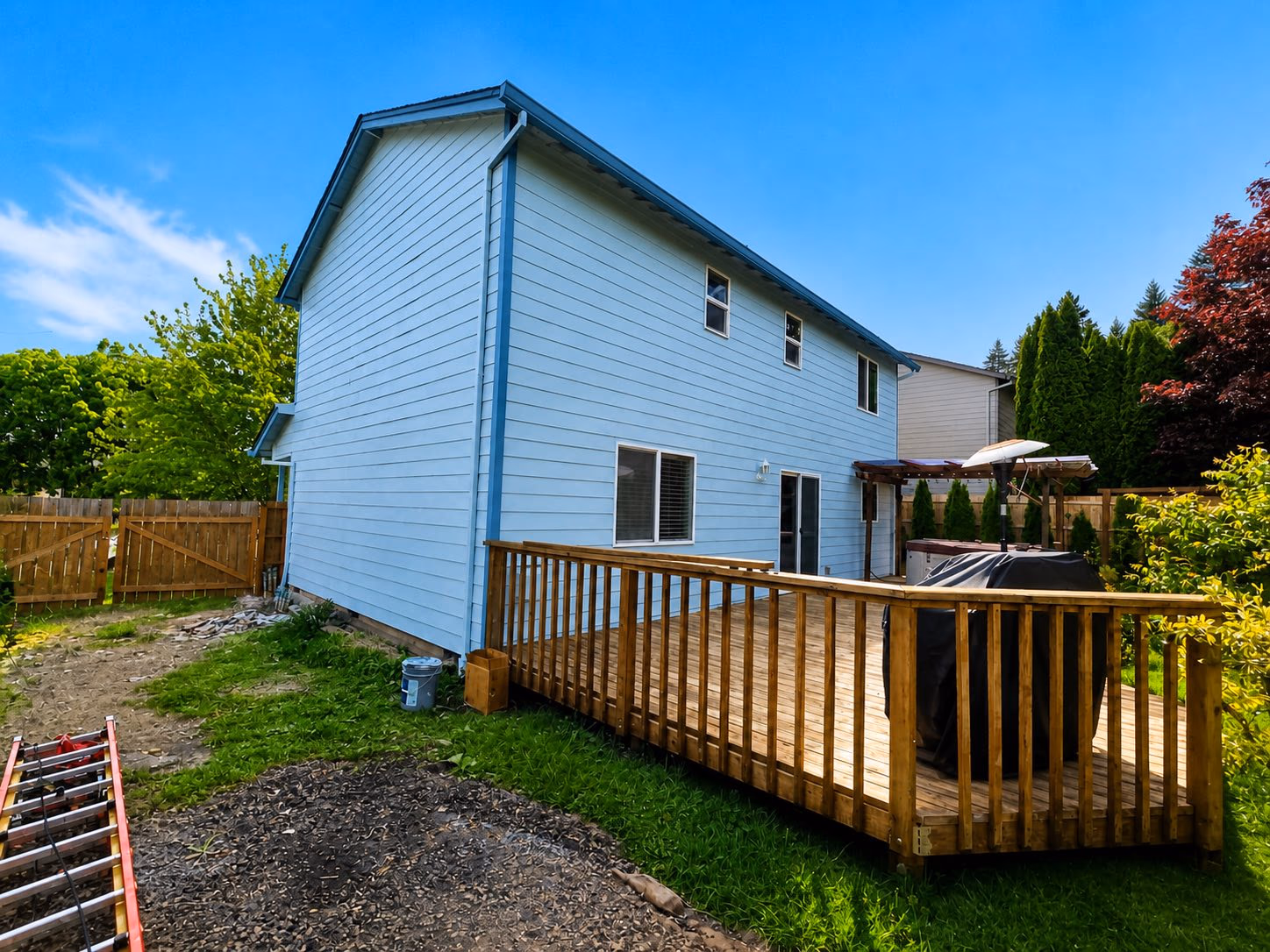 Two-story house with light blue siding, wooden deck, and fenced backyard with green grass and trees.