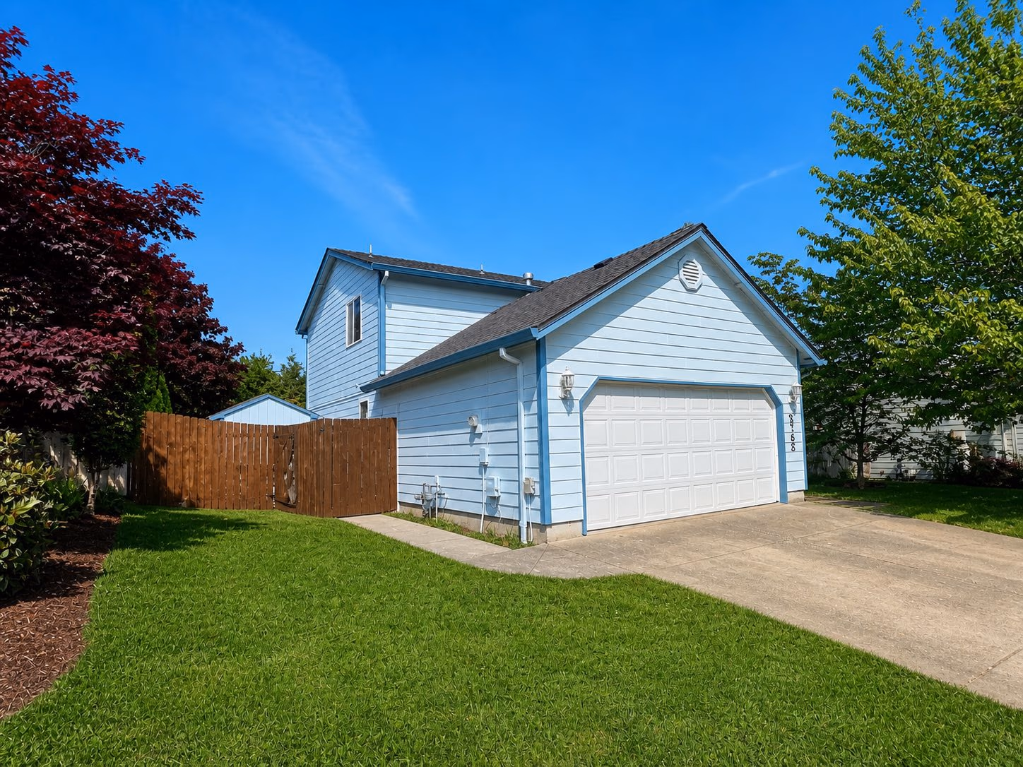 Blue two-story house with white garage door, wooden fence, green lawn, and trees on a sunny day.