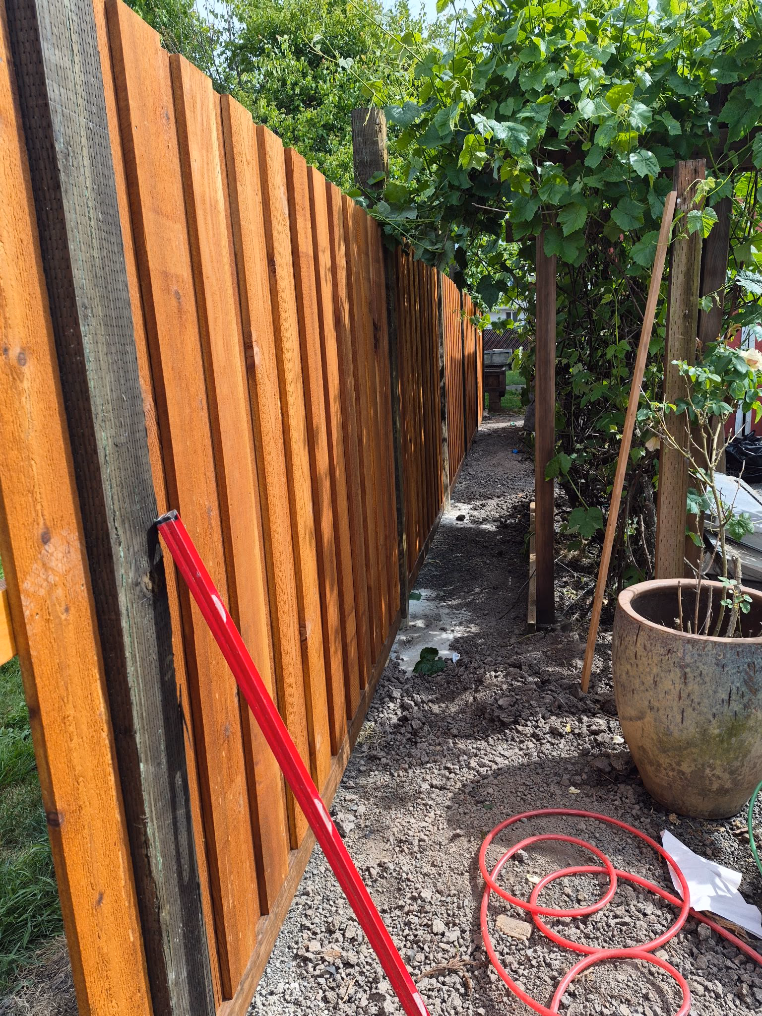 Newly installed wooden fence with red pole and coiled red cable on gravel beside green plants.