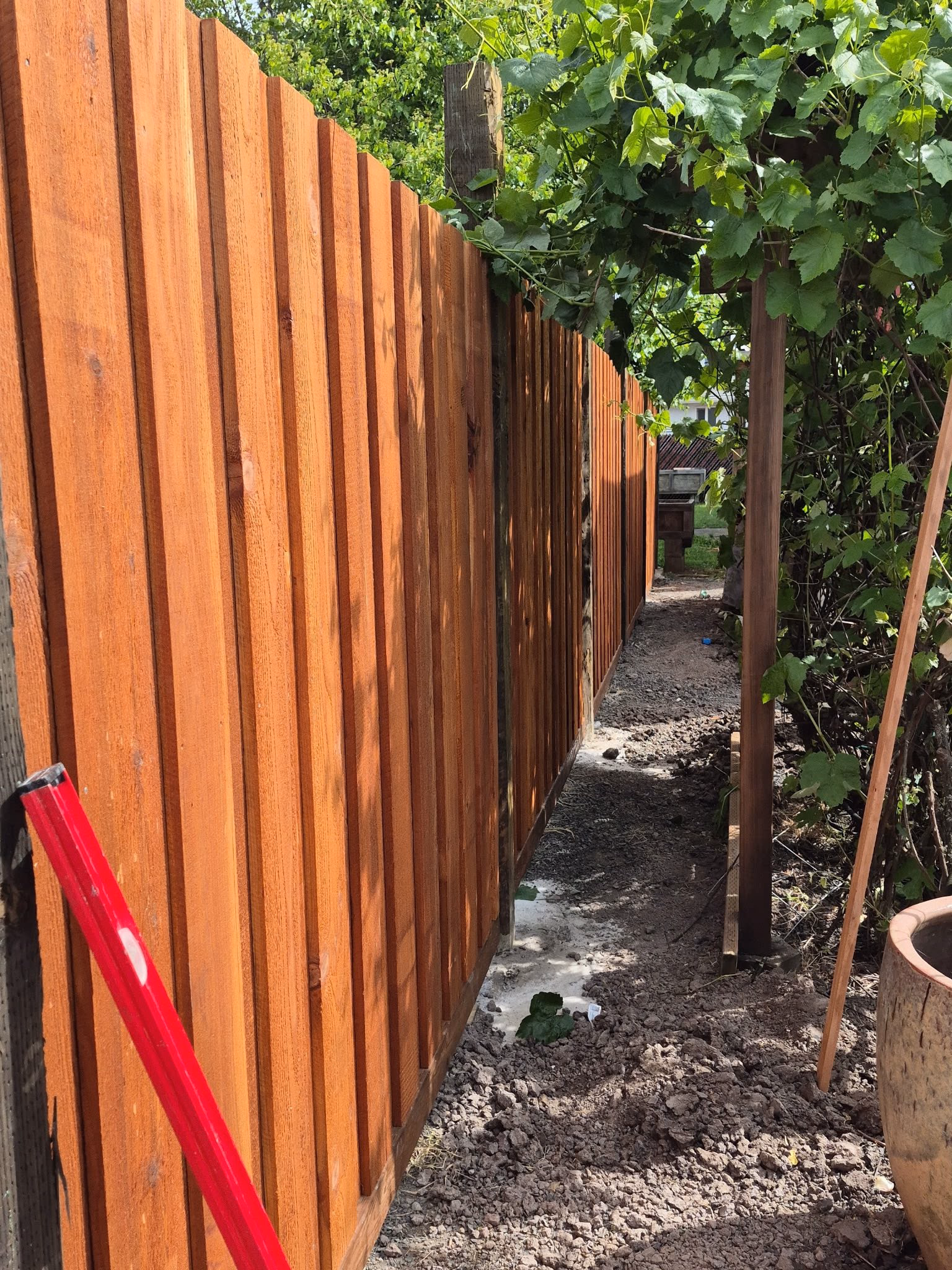 New wooden fence with vertical slats alongside dirt path and green foliage on a sunny day.