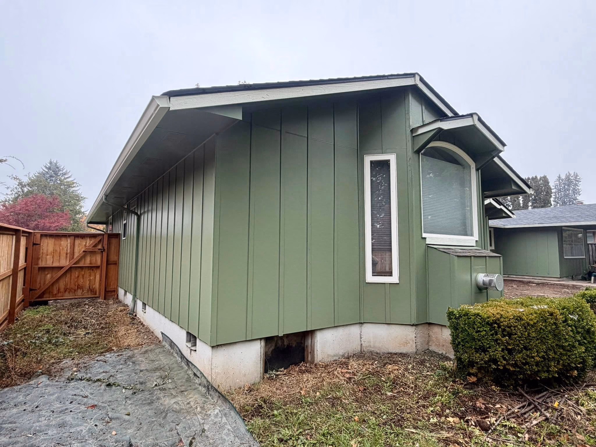 Green house exterior with vertical siding, small windows, a wooden fence, and trimmed shrubbery.