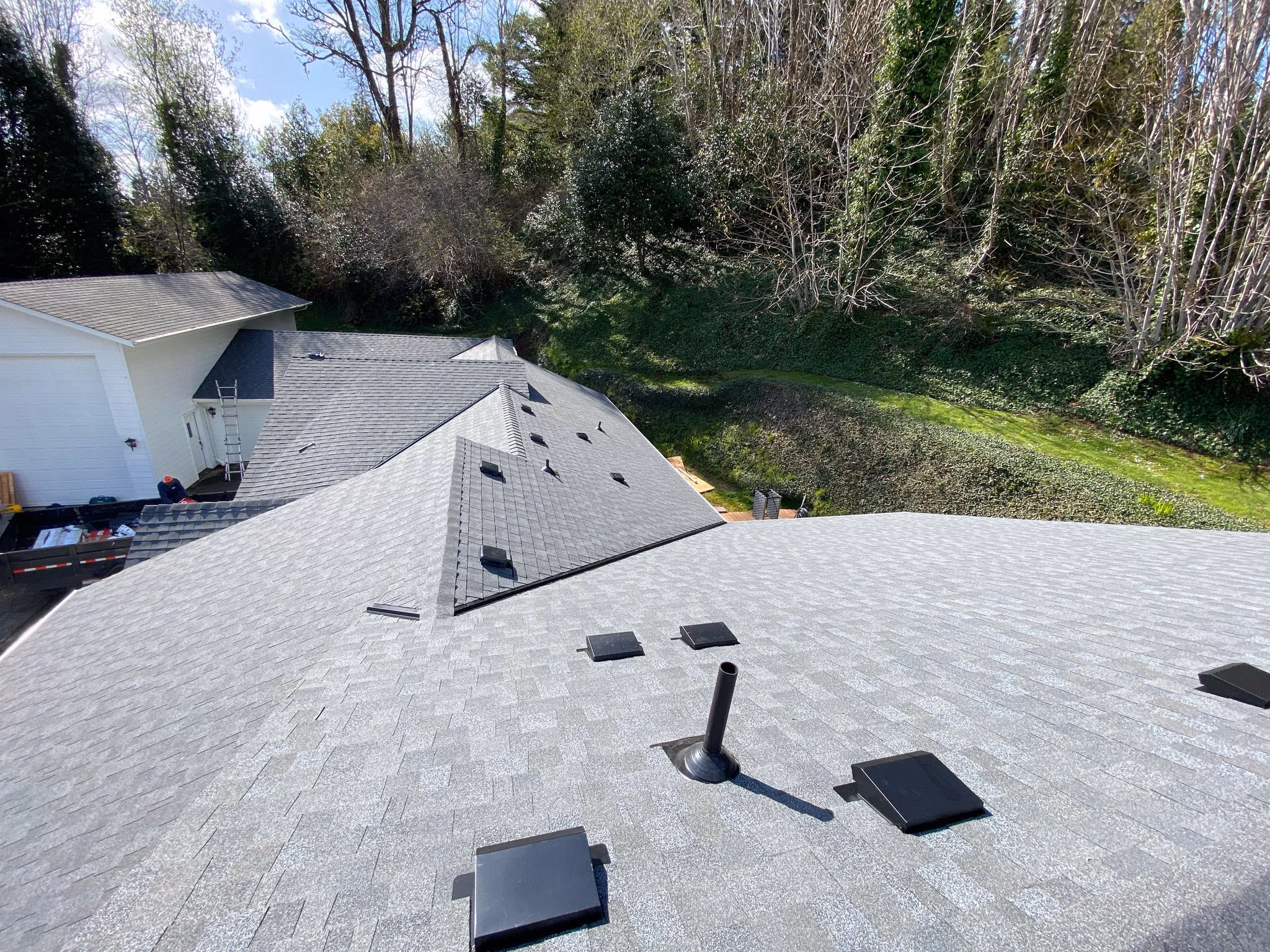 Gray shingled roof with black vents overlooking a green backyard and trees under blue sky.