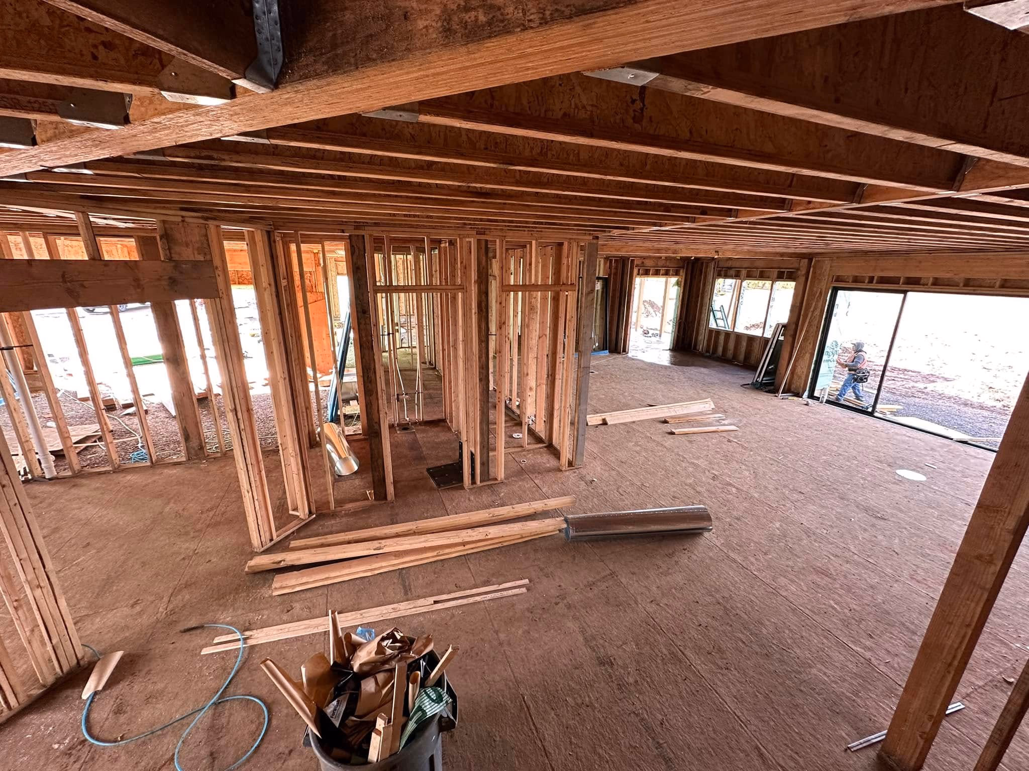 Interior of a house under construction with exposed wooden framing and a worker outside sliding glass doors.