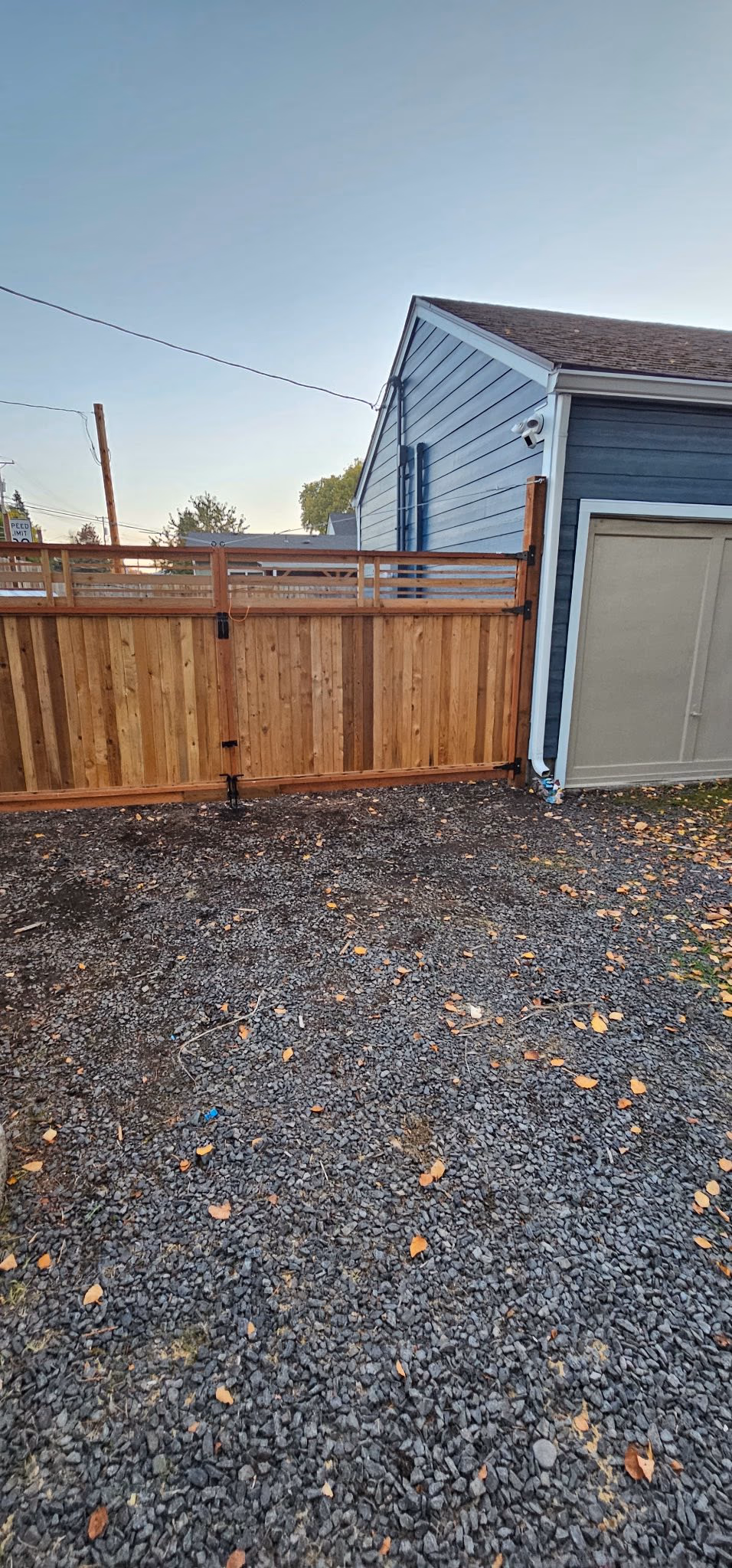 Gravel driveway with autumn leaves leads to wooden fence gate and blue house with security camera in evening.